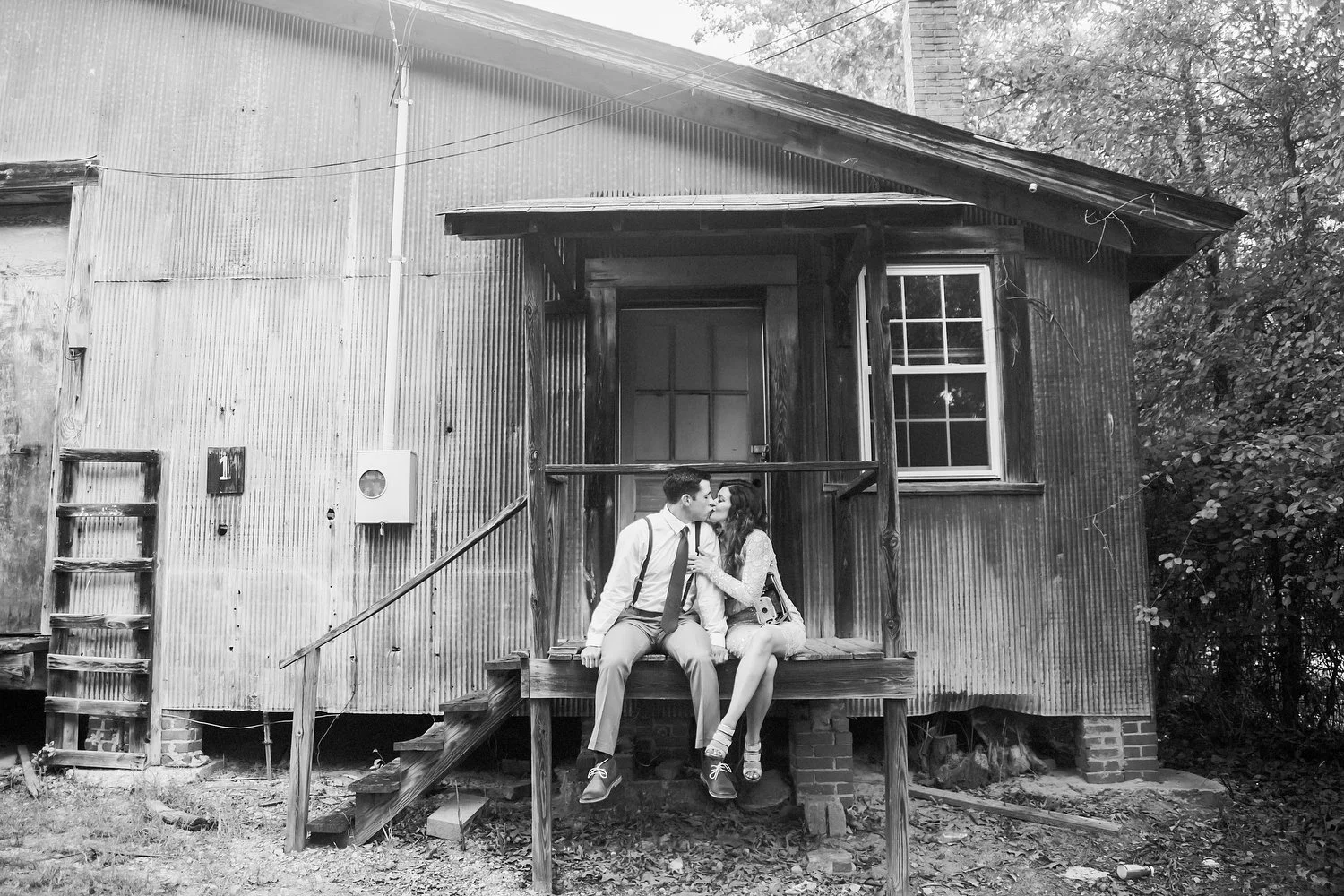 Black and white photo of a couple sitting on a porch landing on the side of an old building that looks like it's built out of a mix of metal panels and wood. They are small in the frame. Her legs are crossed toward him and she leans in with her arm a