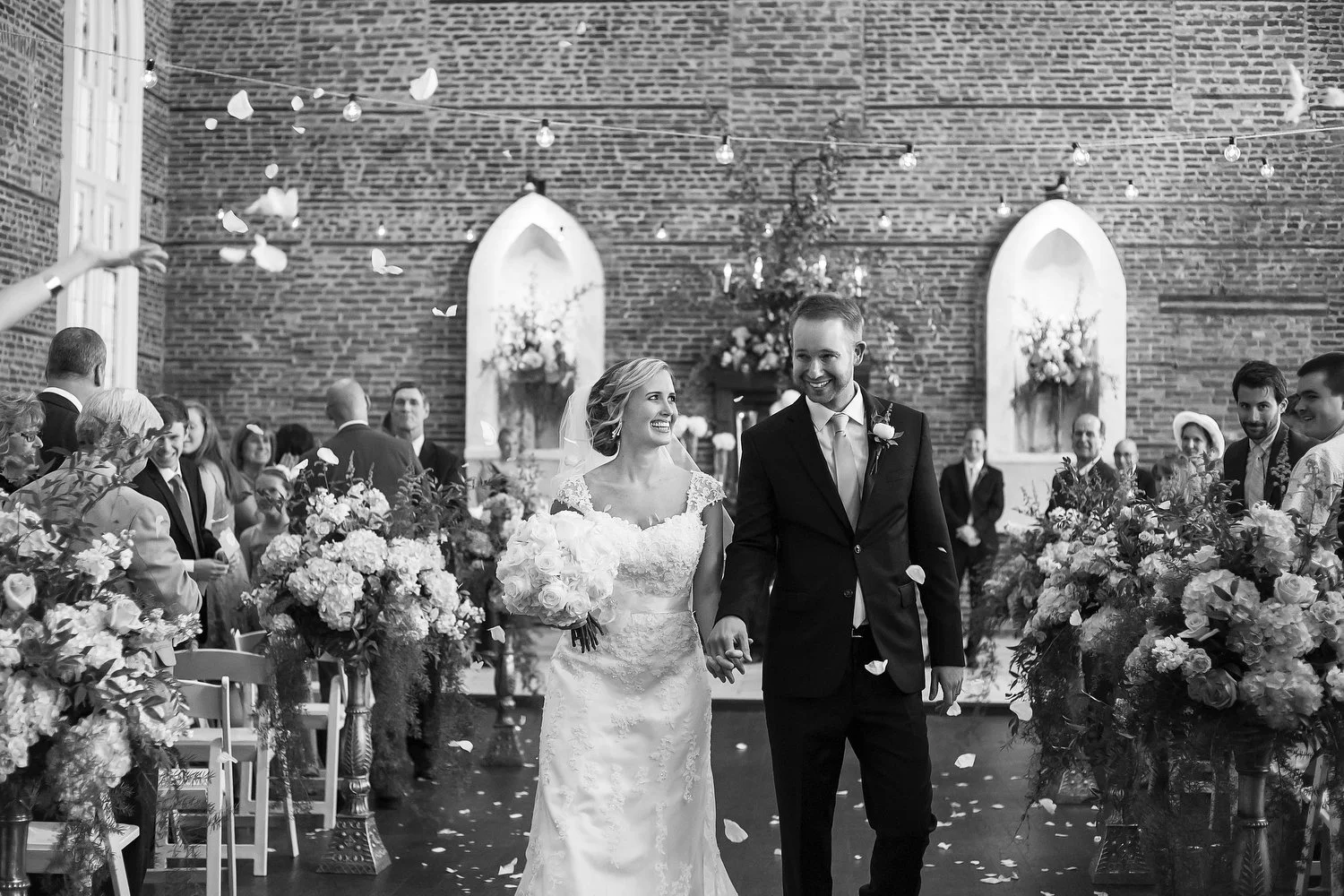 Black and white photo of a bride and groom walking down the aisle in a decorated brick venue, with guests on either side and flower arrangements.