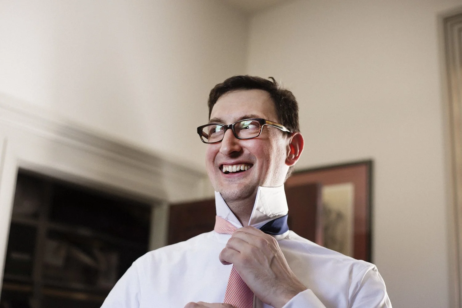 A smiling groom with glasses adjusting his light red tie in a well-lit room.