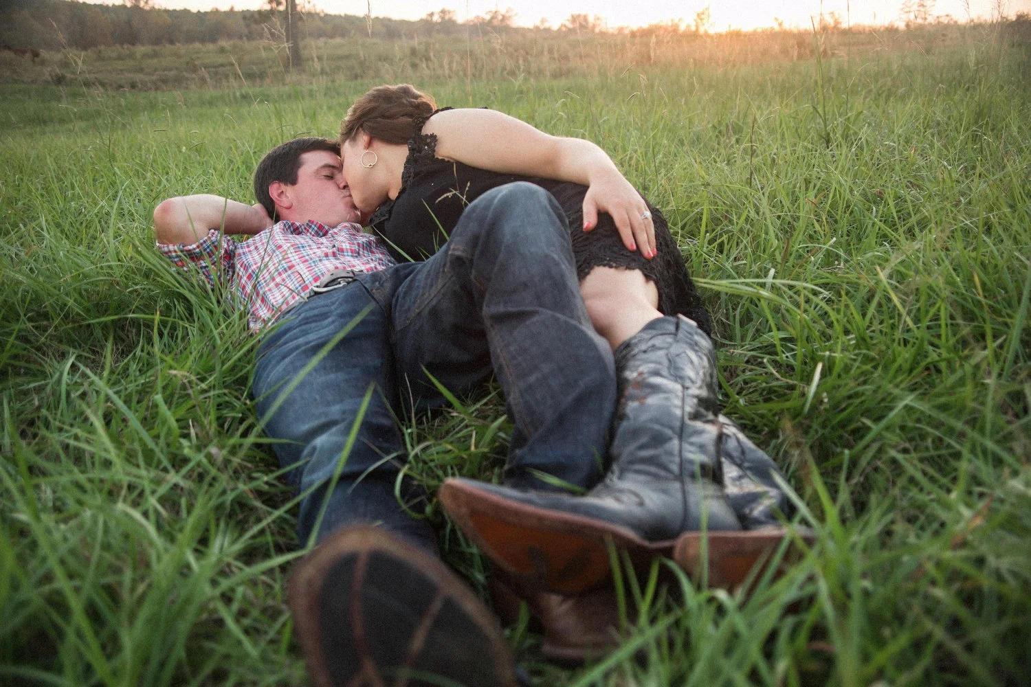 A man and a woman both wearing cowboy boots are lying in a field of green grass. He is on his back with his left leg bent and his right arm behind his head. He is wearing a checkered button-up shirt and jeans. She is on her side and leaning in to kis