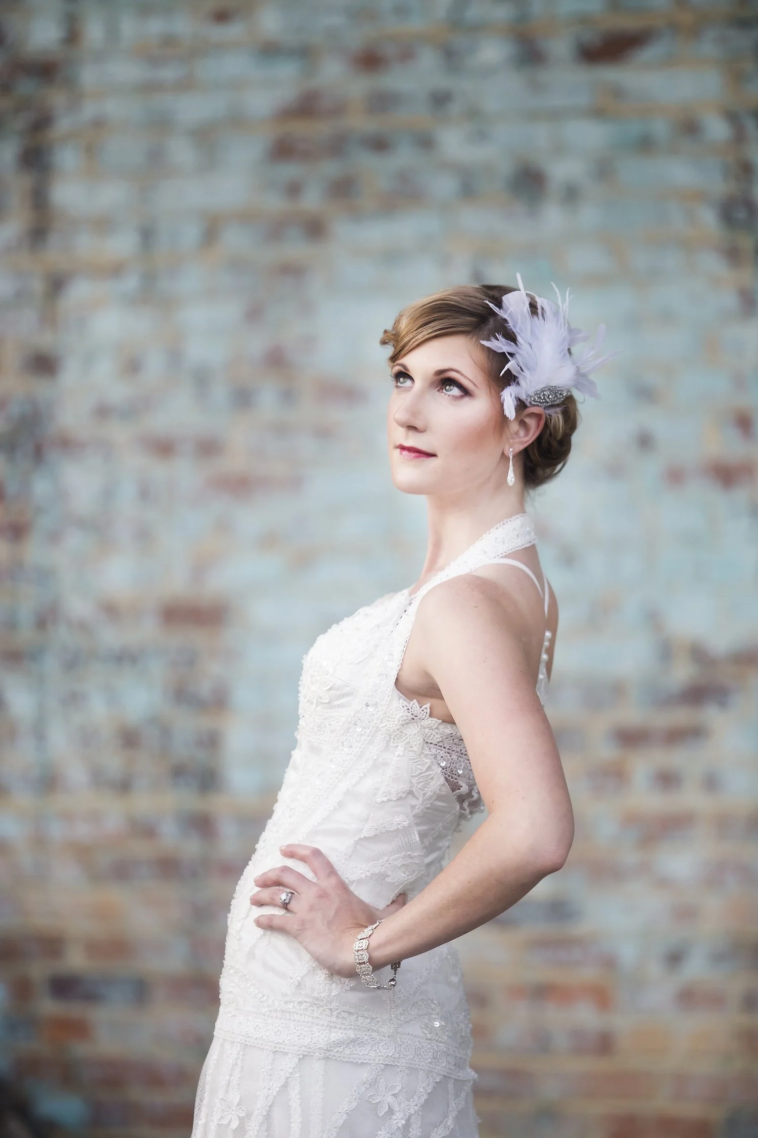 A woman in a white lace wedding dress with a feather headpiece, standing with her hand on her hip against a textured red and teal brick wall, looking pensively to the side.