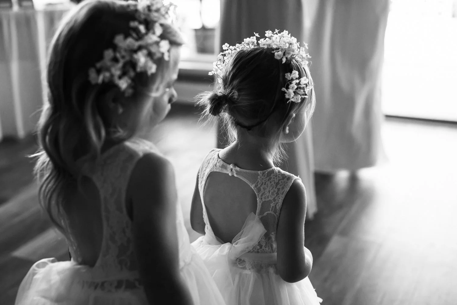 Black and white image of two young girls dressed in lace and tulle, wearing flower crowns, standing indoors near a window with light streaming in.