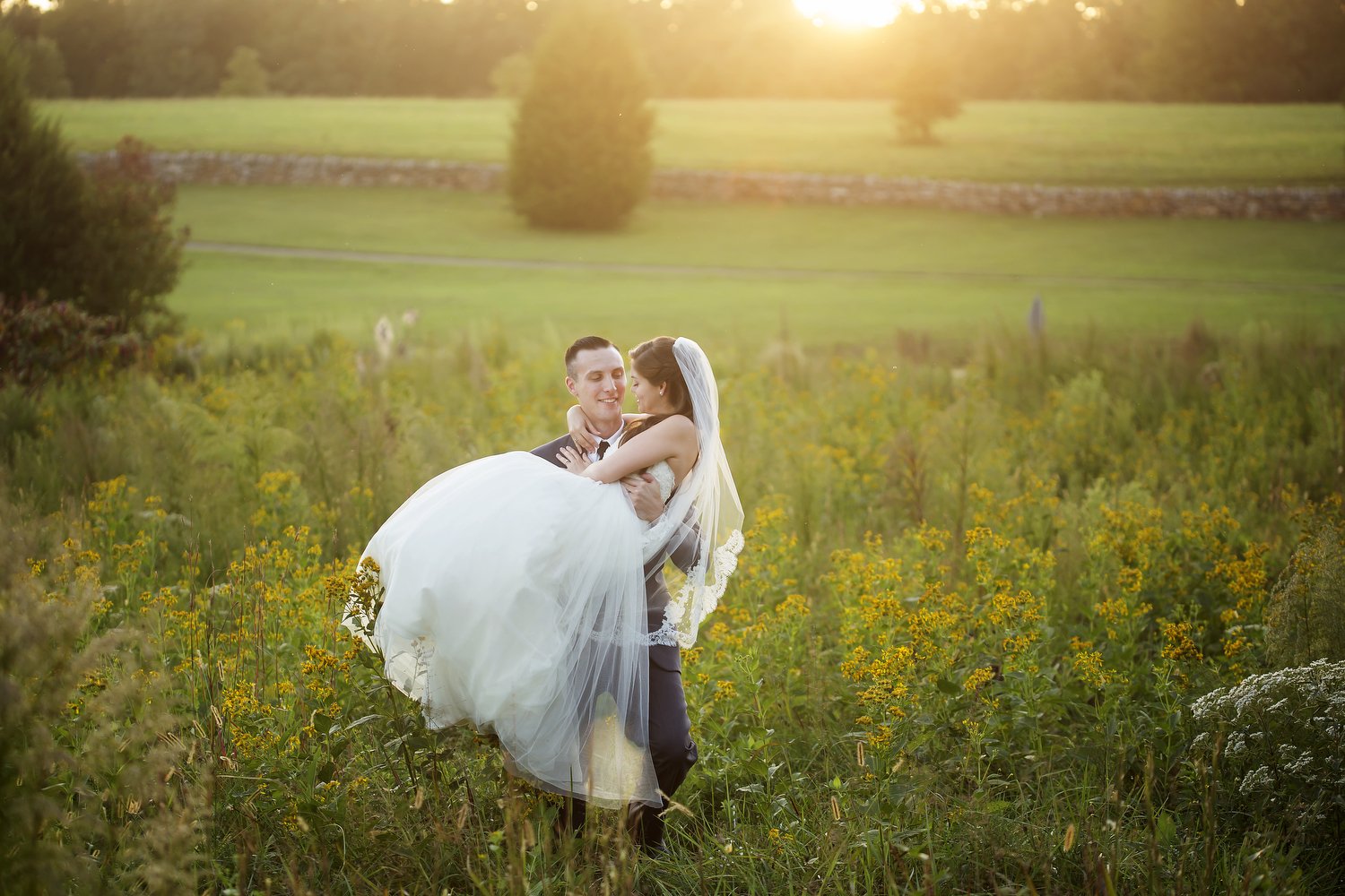 A groom lifts his bride and carries her across a green field with yellow flowers at sunset with a glow of light behind them. She is wearing a flowy white dress and a long veil.