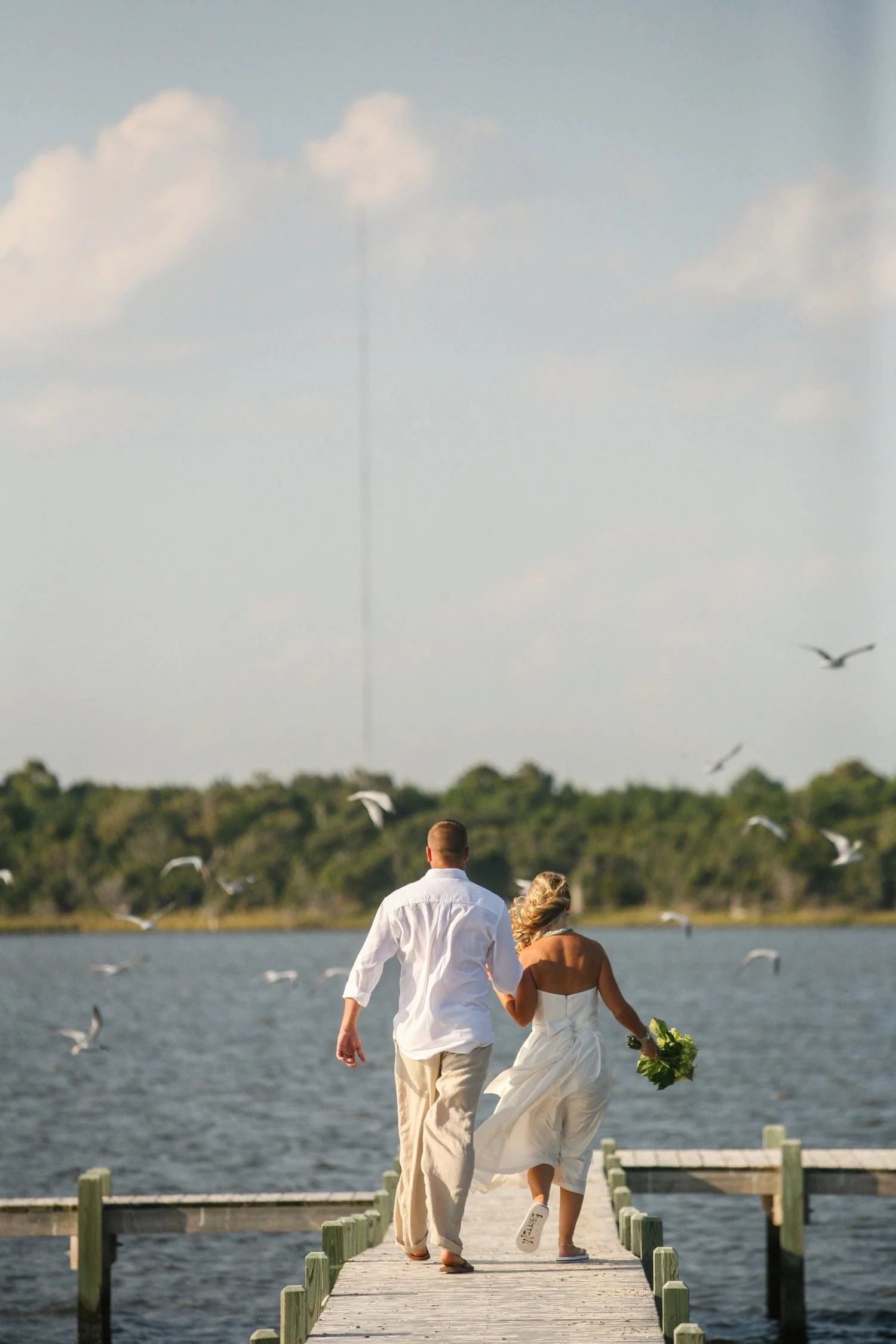 A bride and groom are seen from behind as they walk down a wooden dock toward the water. She is wearing a tea-length white strapless dress and holding a bouquet in her right hand. He has on khaki pants and a white dress shirt. Seagulls are flying up 