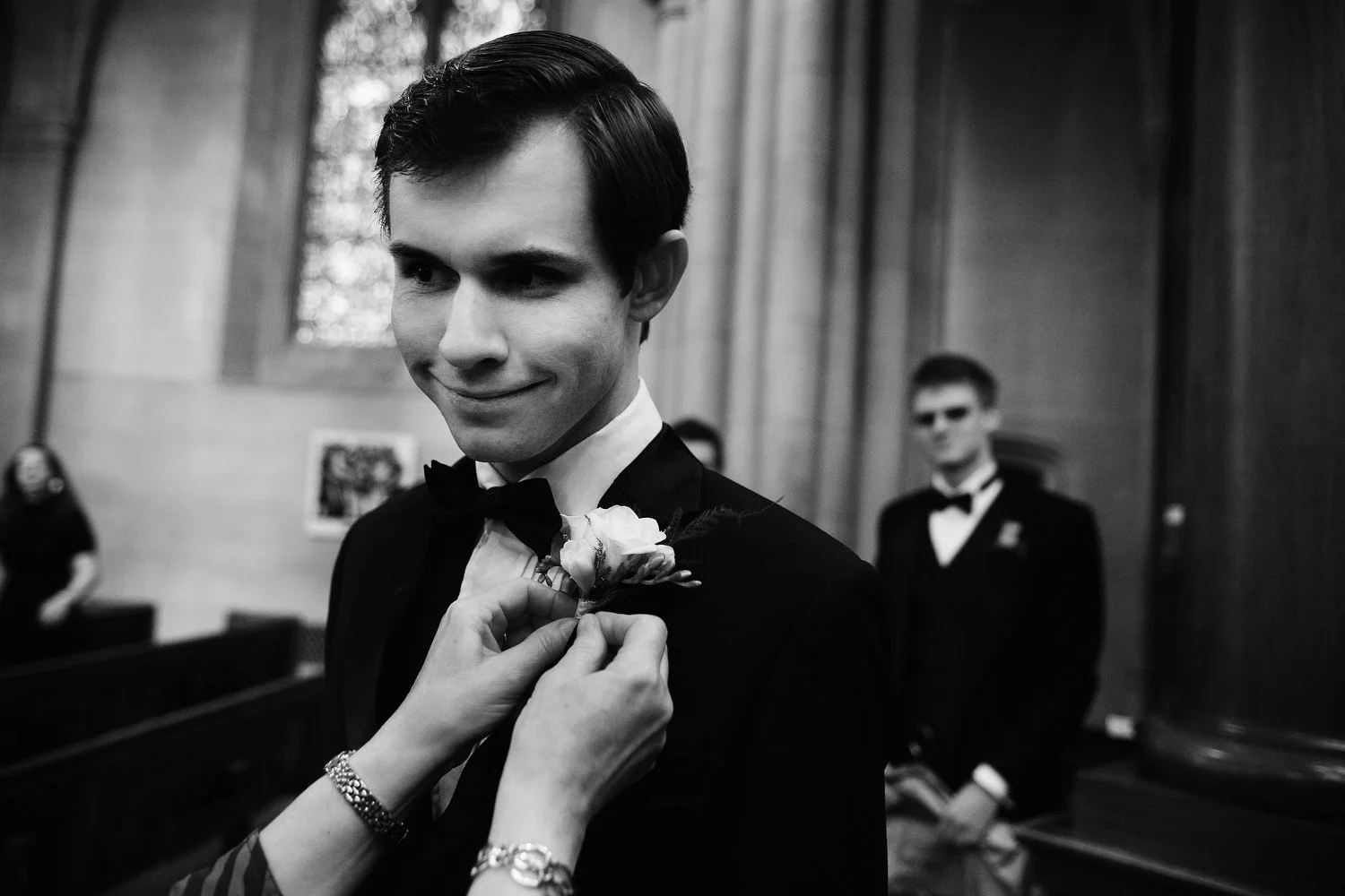 Black and white photo of a young man in a tuxedo, smiling as a woman pins a collar boutonniere on him before his wedding ceremony in Duke Chapel. You can see stained glass windows behind him.