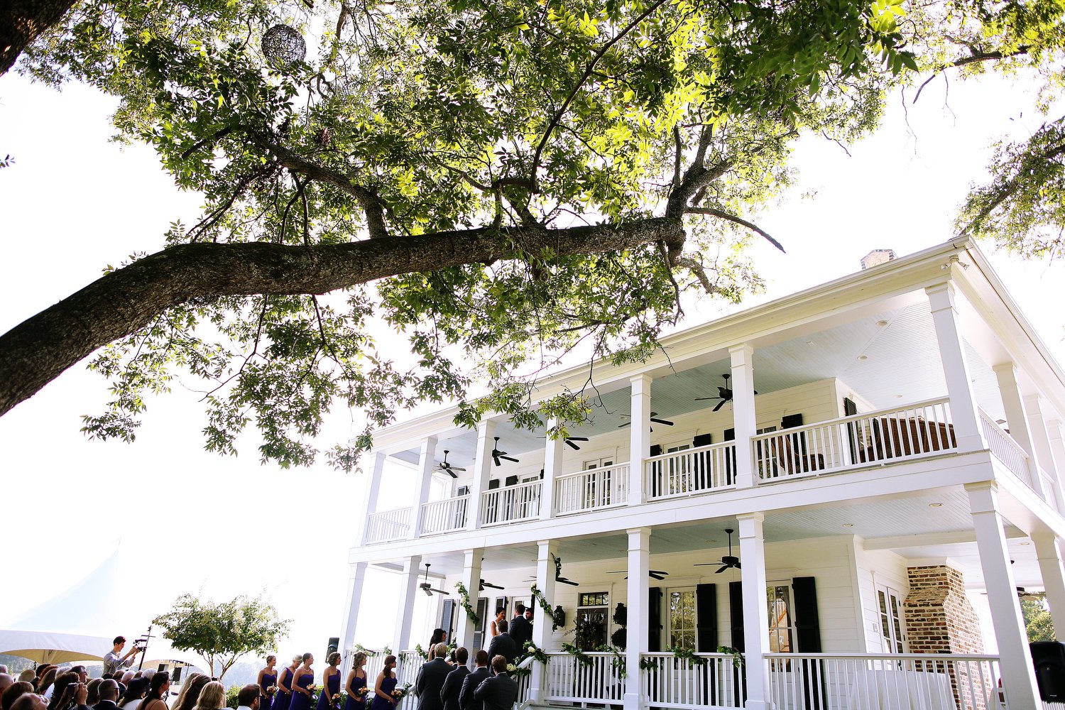 A wedding ceremony taking place outdoors in front of a large white house with multiple porches. Bridesmaids in blue dresses and groomsmen in dark suits are standing in a line, facing a couple who are exchanging vows. A large tree with green leaves is
