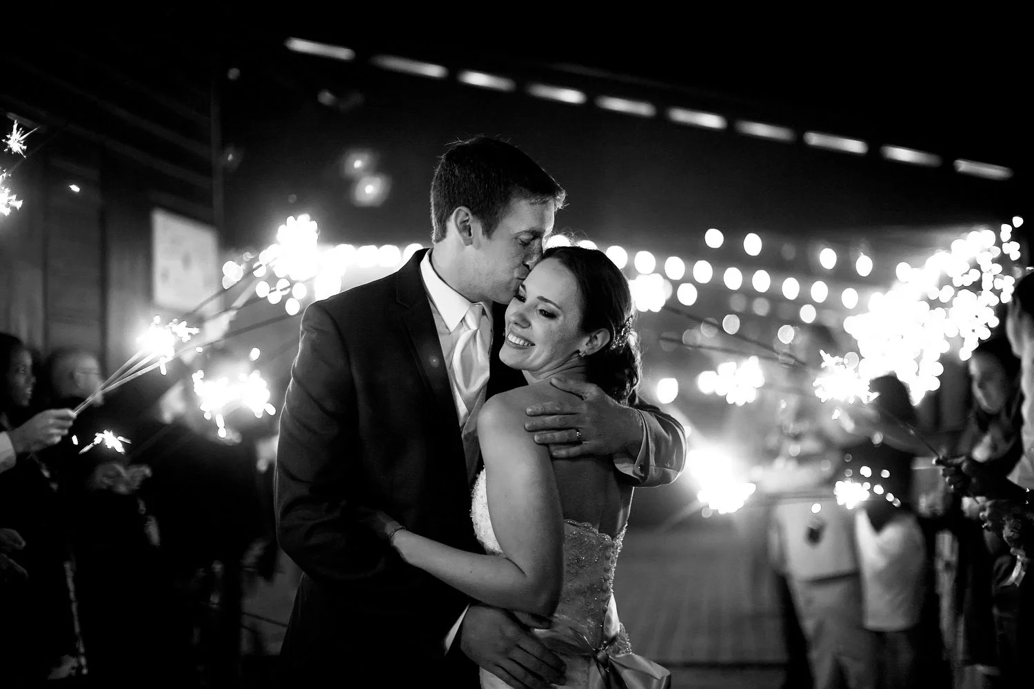 Black and white photo of a happy couple hugging as they exit their wedding reception, surrounded by sparklers held by guests outside a decorated barn venue.