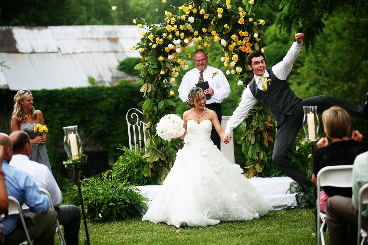 A joyful wedding ceremony outdoors with a bride in a white strapless wedding gown, a groom in a black vest and yellow tie, and a officiant under a floral arch adorned with yellow roses. Guests sit on white chairs. The groom is excitedly leaping in th