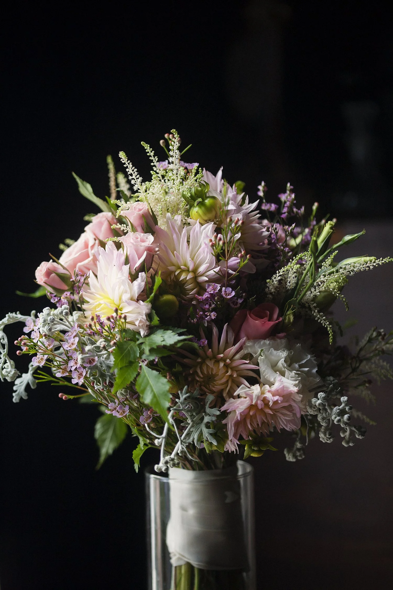 Vertical image of a bridal bouquet in a glass jar of water. The bouquet is wrapped in a white silk material. Flowers are a mixture of pink, purple, and white and greenery is mixed in. 