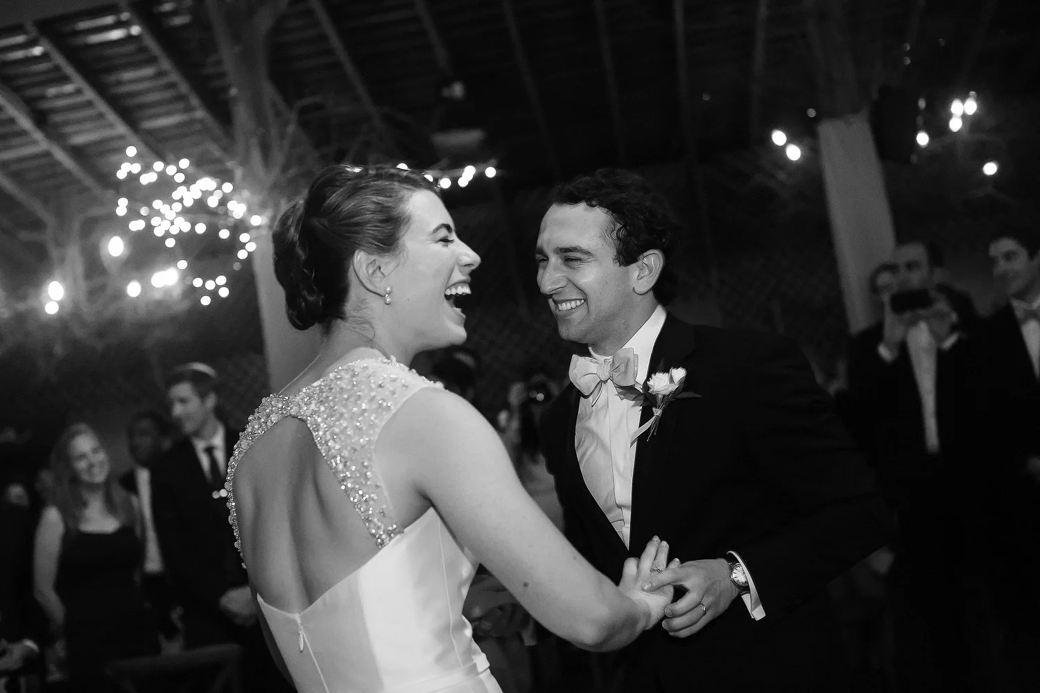 Black and white photo of a bride and groom dancing and smiling at their wedding reception in a decorated venue with string lights, surrounded by guests.