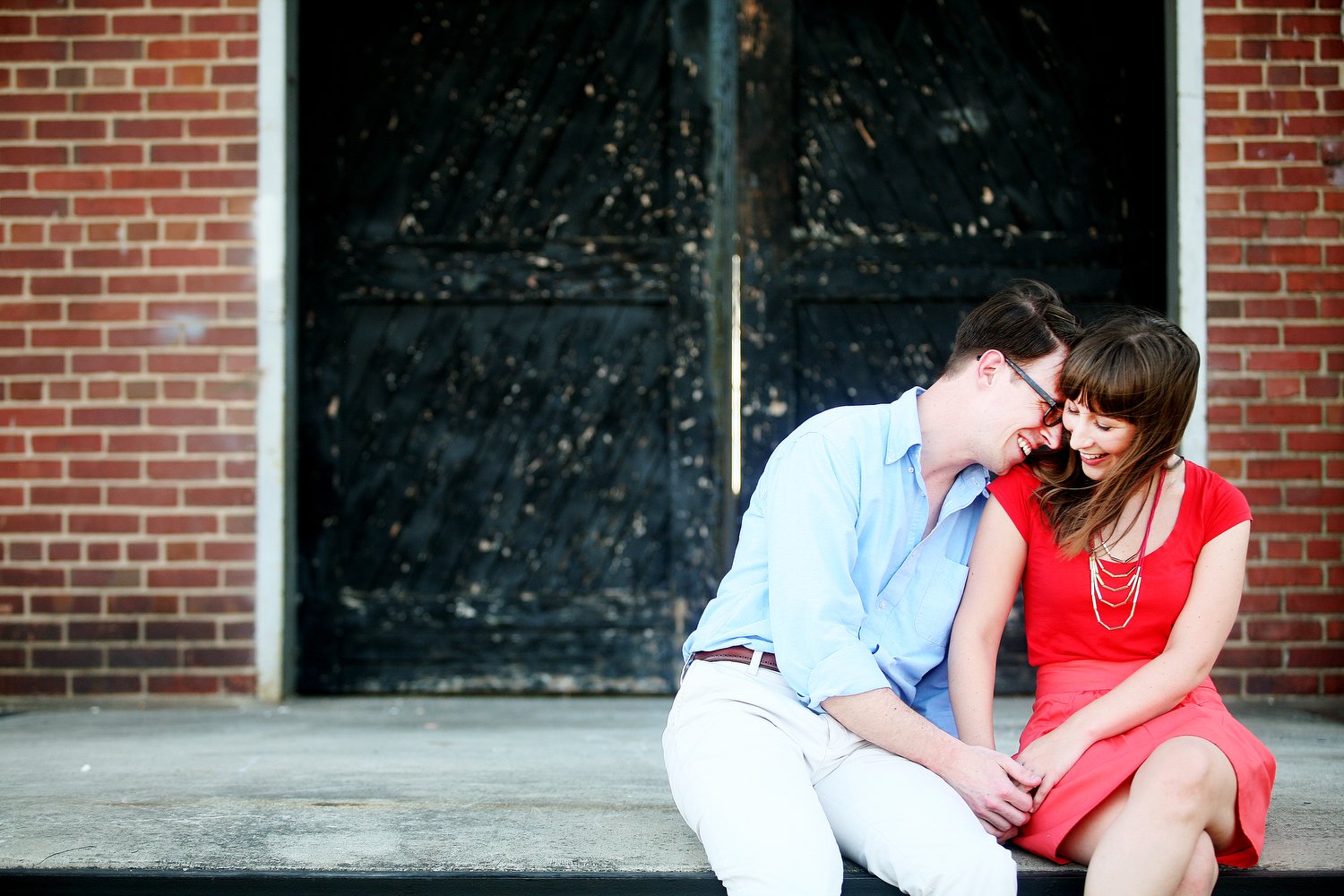 A couple sits on the edge of a concrete landing. There are double black doors behind them and walls of red brick on either side of the doors. She has on a red dress and necklace and holds his hand. Her legs are crossed toward him. He leans his head a