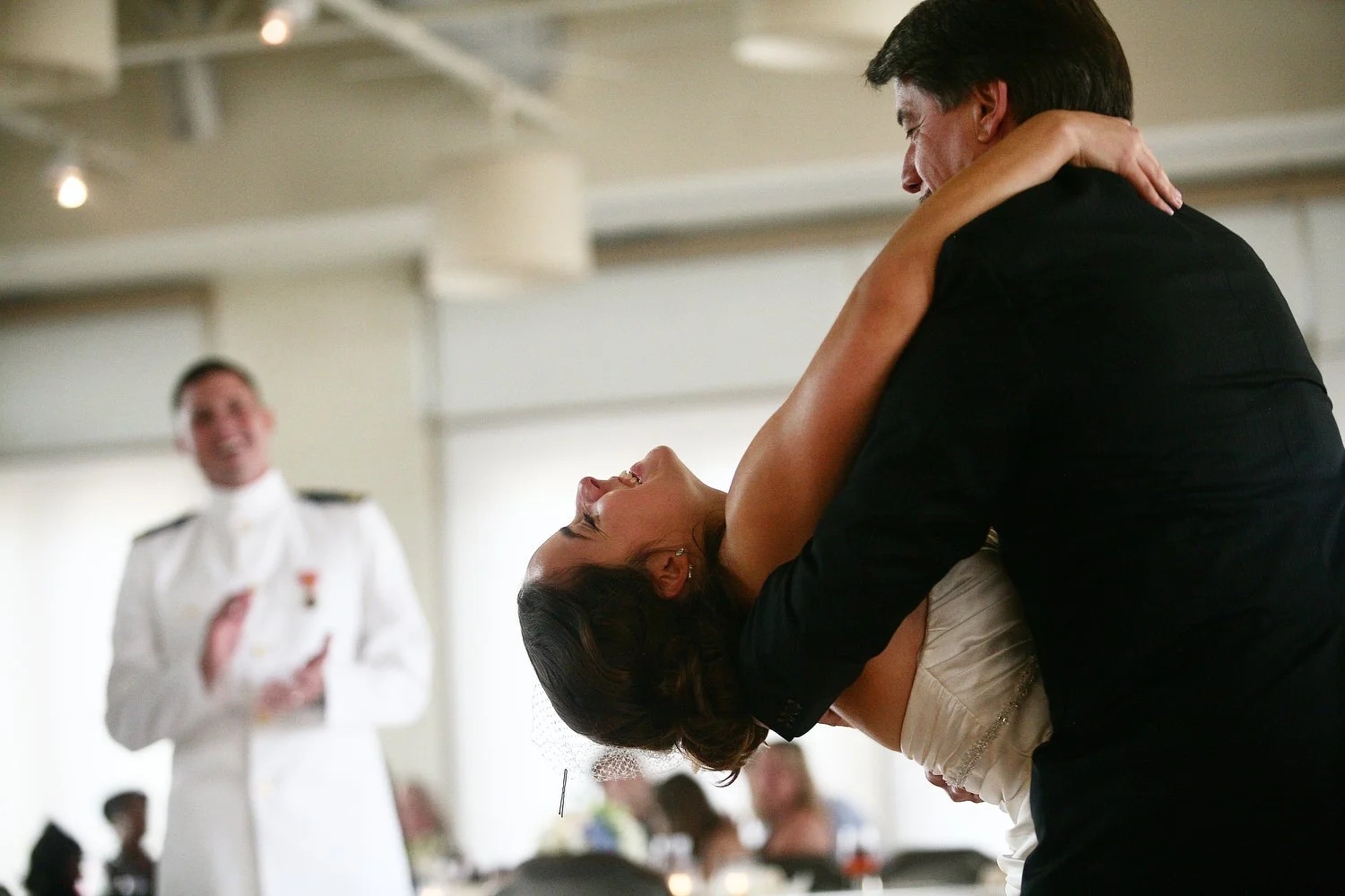 A bride and her father dance at her wedding reception. Her dad bends her back to dip her as the groom, in a military uniform, claps and smiles in the background.