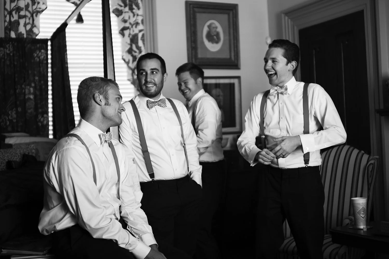Black and white photo of four men dressed in white dress shirts, suspenders, and bow ties, smiling and talking in a room with framed pictures, floral curtains and a canopy bed.
