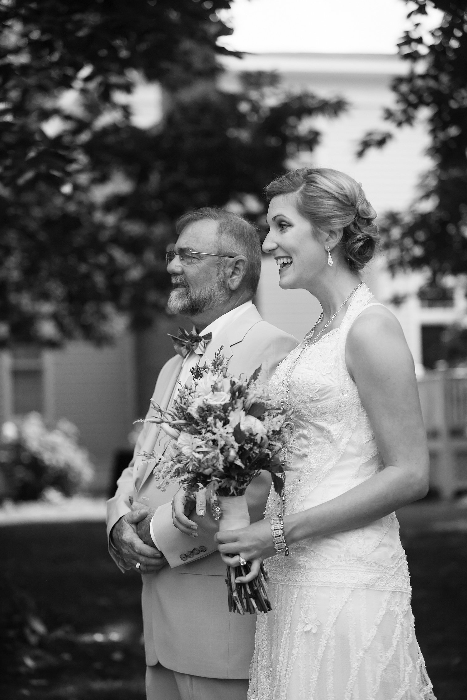 Black and white photo of a bride in a lace wedding dress holding a bouquet, standing next to an older man in a suit with a bow tie, outdoors with trees and a house in the background.