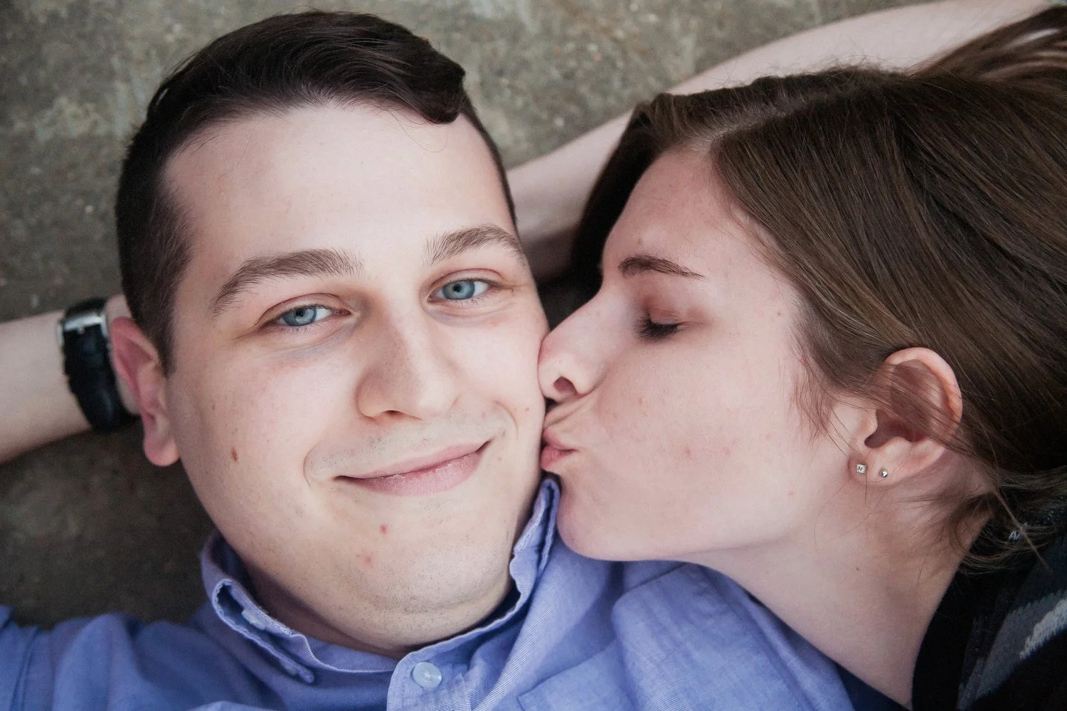 Close up photo taken from above of a woman with brown hair and eyes closed lying on a man's left shoulder and kissing him on the cheek. He is wearing a blue dress shirt and is smiling up at the camera with his arms behind his head on the ground.