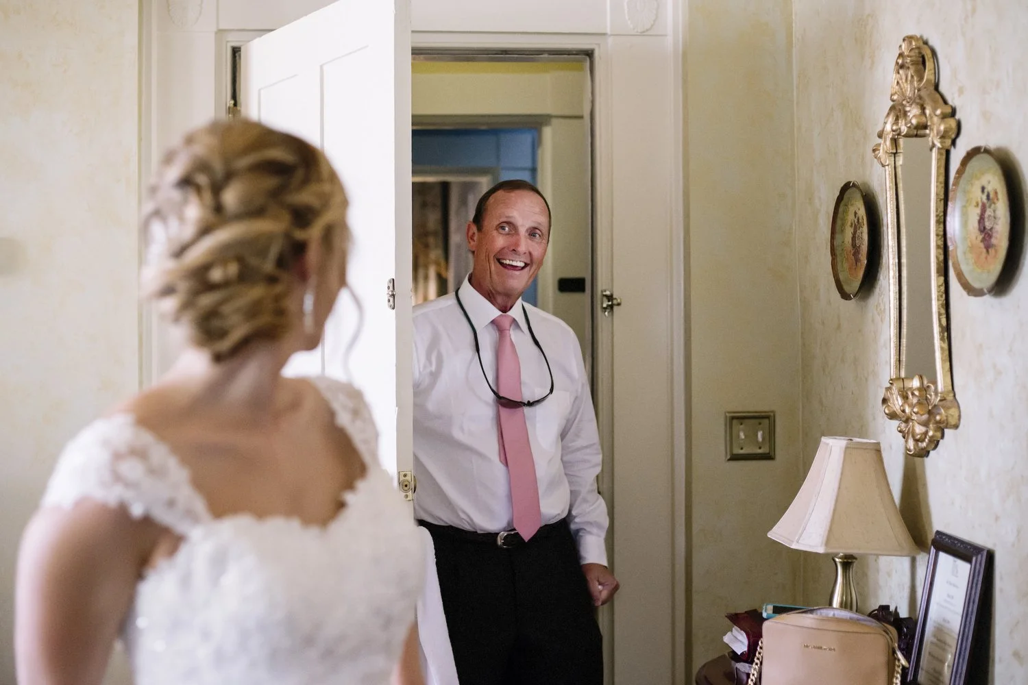 A smiling man in a white dress shirt, pink tie, and glasses hanging around his neck, stands in a doorway and looks at a woman in a white wedding dress, seen from behind, inside a decorated room. The bride is looking back at her dad as he comes in the