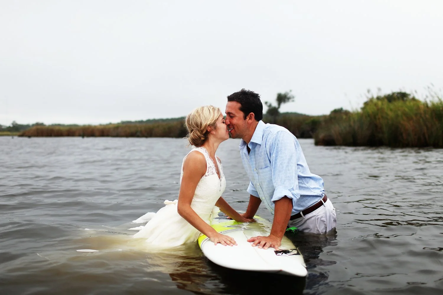 A couple stands in the water in a marshy area at the coast. Their hands are placed on a surfboard that floats between them as they lean in and kiss above it. She has on a wedding dress and her hair is pulled up in a messy bun. He has on khaki pants a