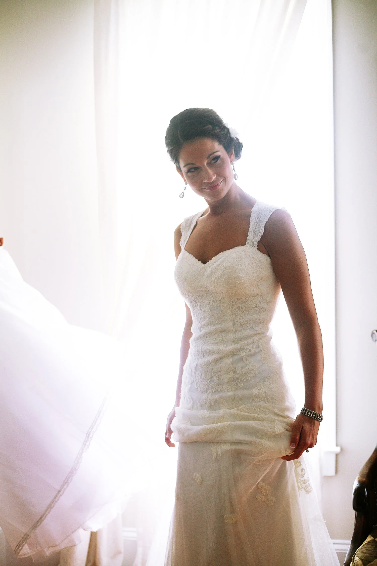 A bride in a wedding dress standing near a bright window with sheer curtains, smiling softly.