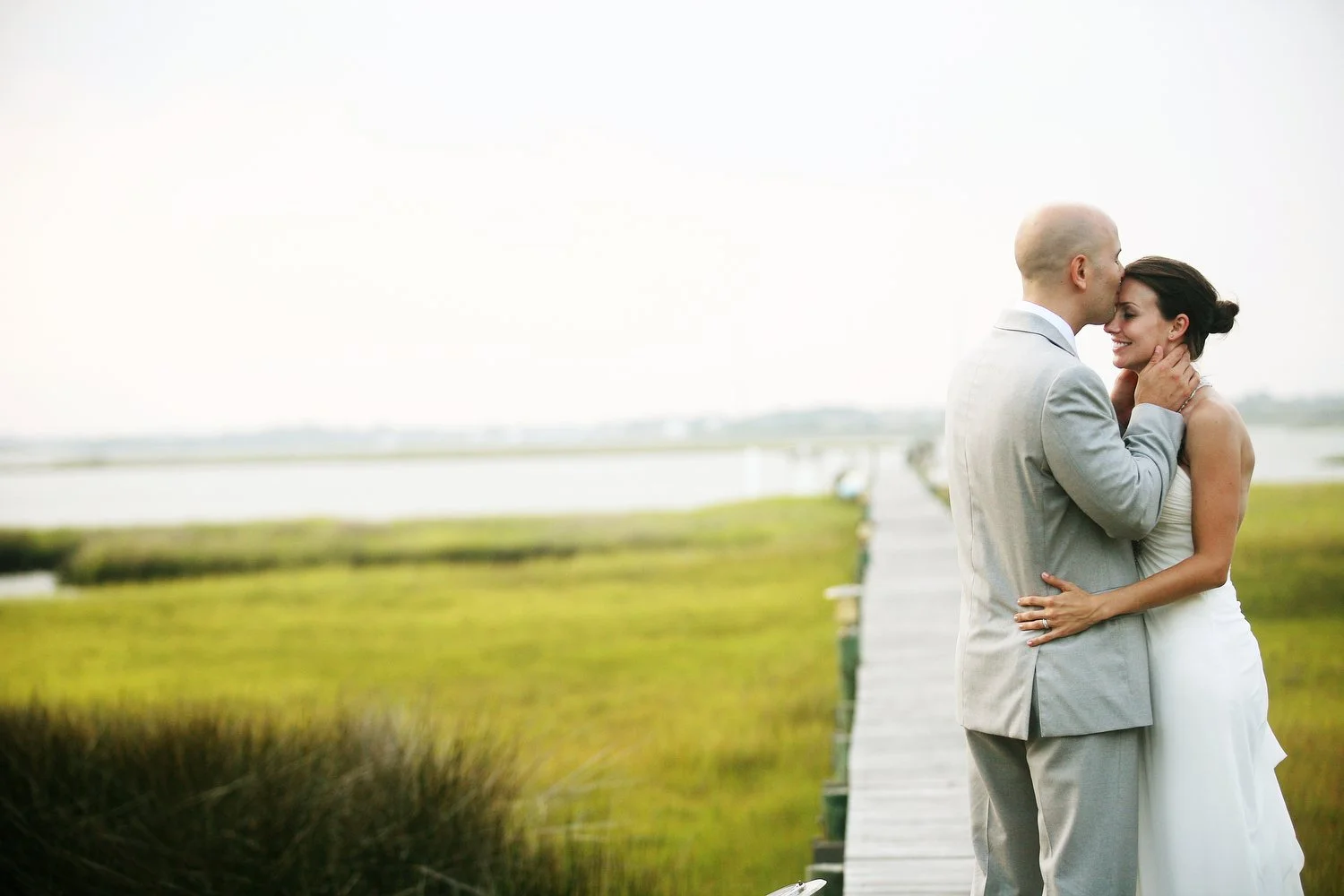 A bride and groom embrace on a wooden dock outdoors, with water and marsh grasses in the background. He kisses her forehead as she leans into him.