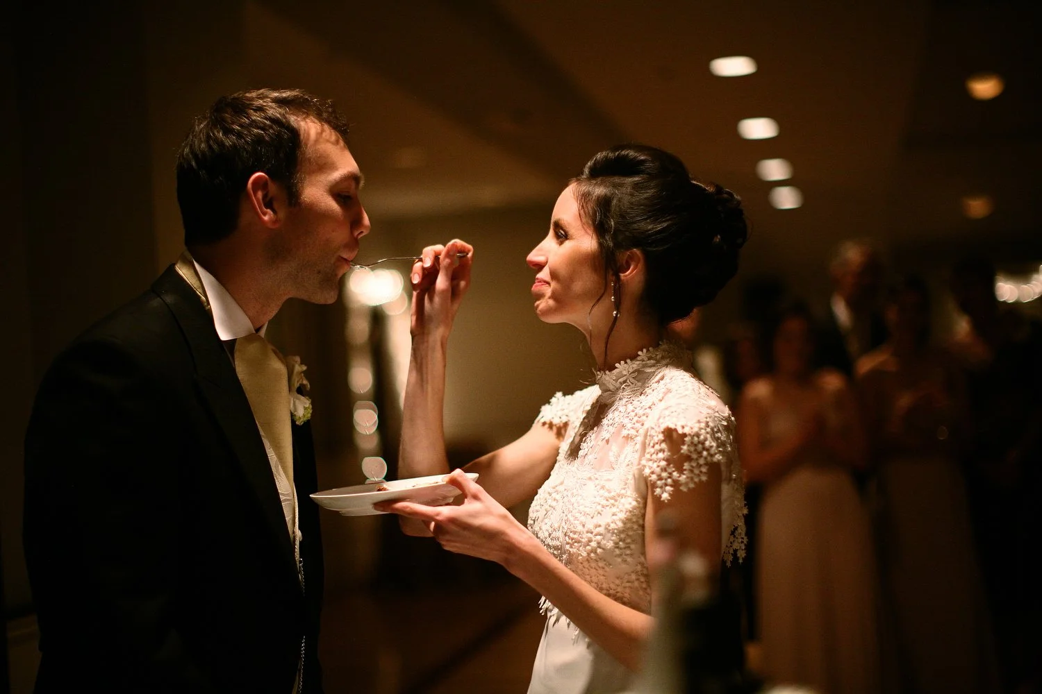 A bride feeds cake to her groom at their wedding reception, with guests in the background.