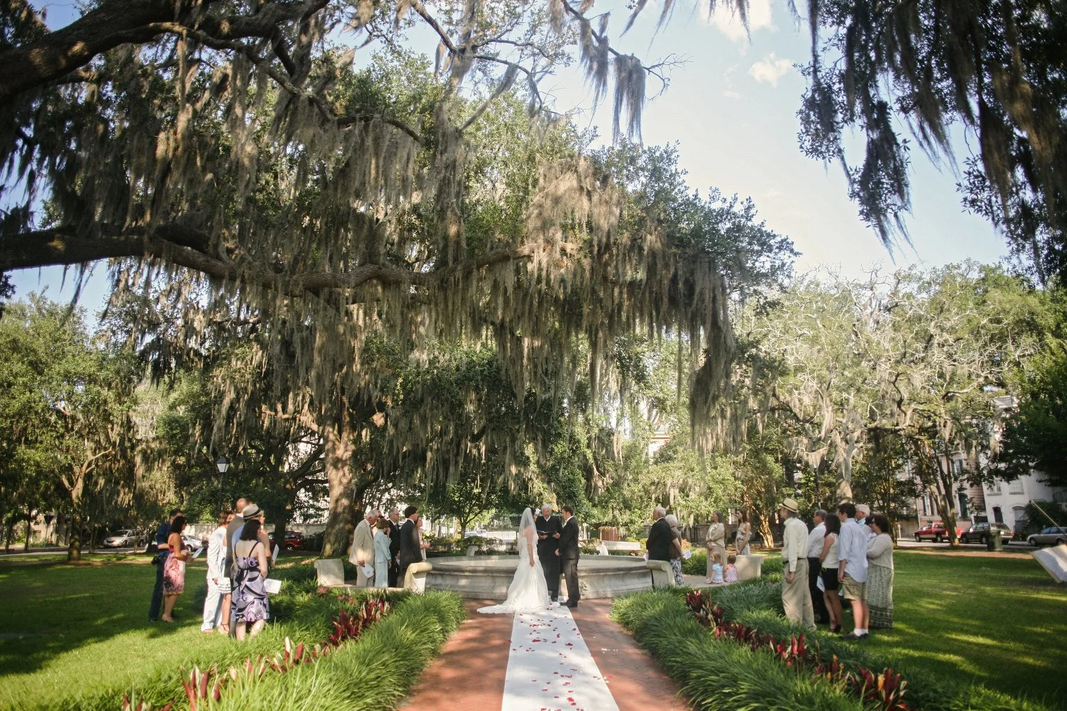 Wedding ceremony taking place outdoors under large trees with Spanish moss in a square in Savannah, with guests gathered around as the bride and groom exchange vows.