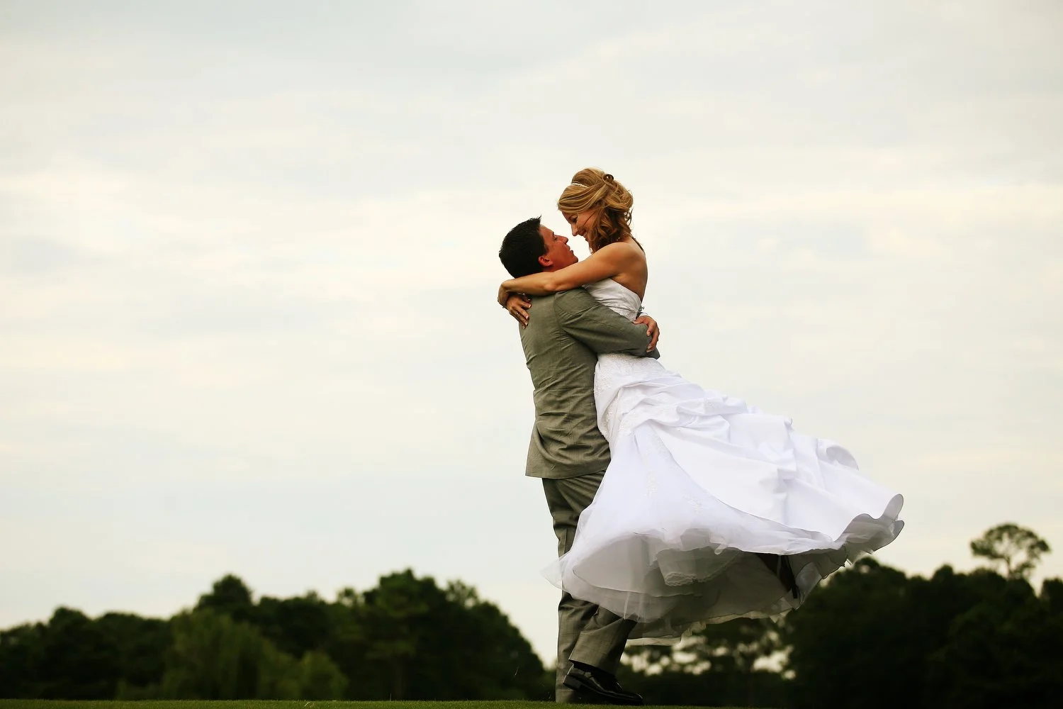 A groom in a grey suit is lifting his bride in a white bunched up wedding dress, with an outdoor landscape in the background.