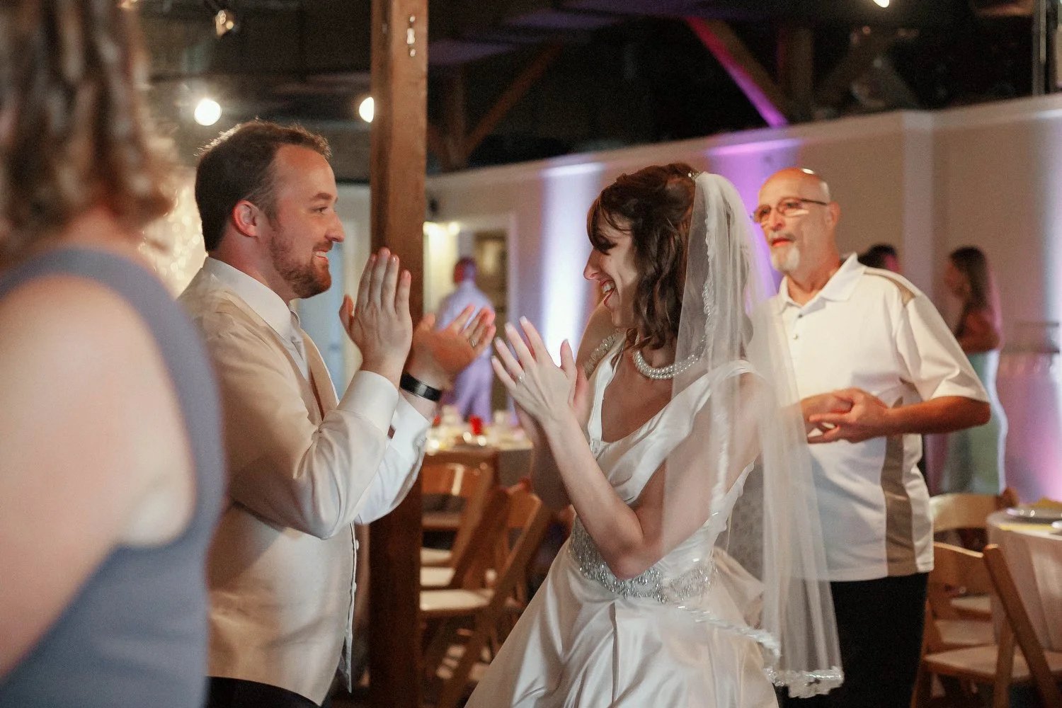 A bride and groom sharing a joyful moment, smiling at each other with their hands pressed together, during a wedding reception. An older man with glasses and a beard stands in the background, smiling and clapping.