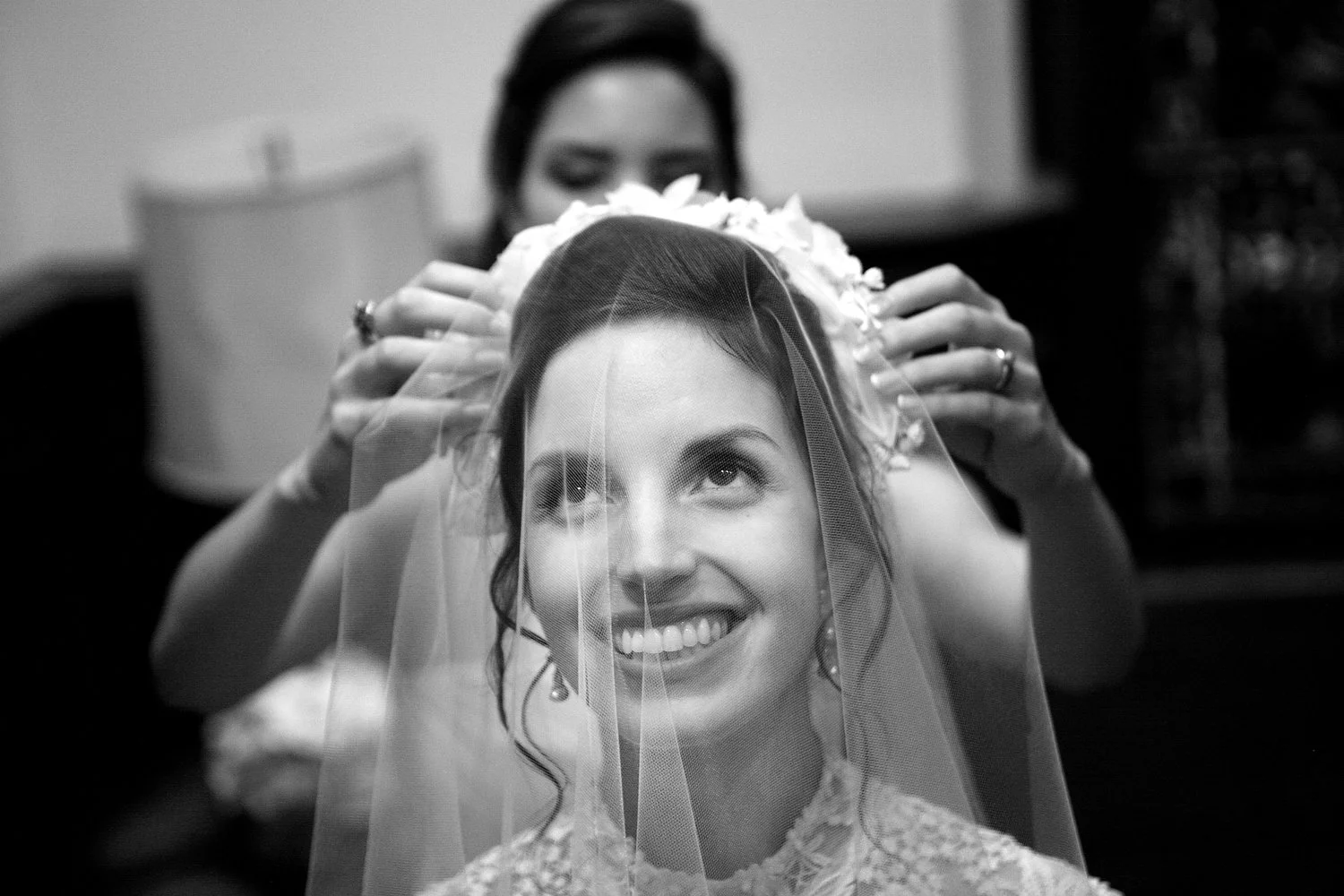 Black and white close up of a bride smiling as a woman places a veil on her head during her wedding day.