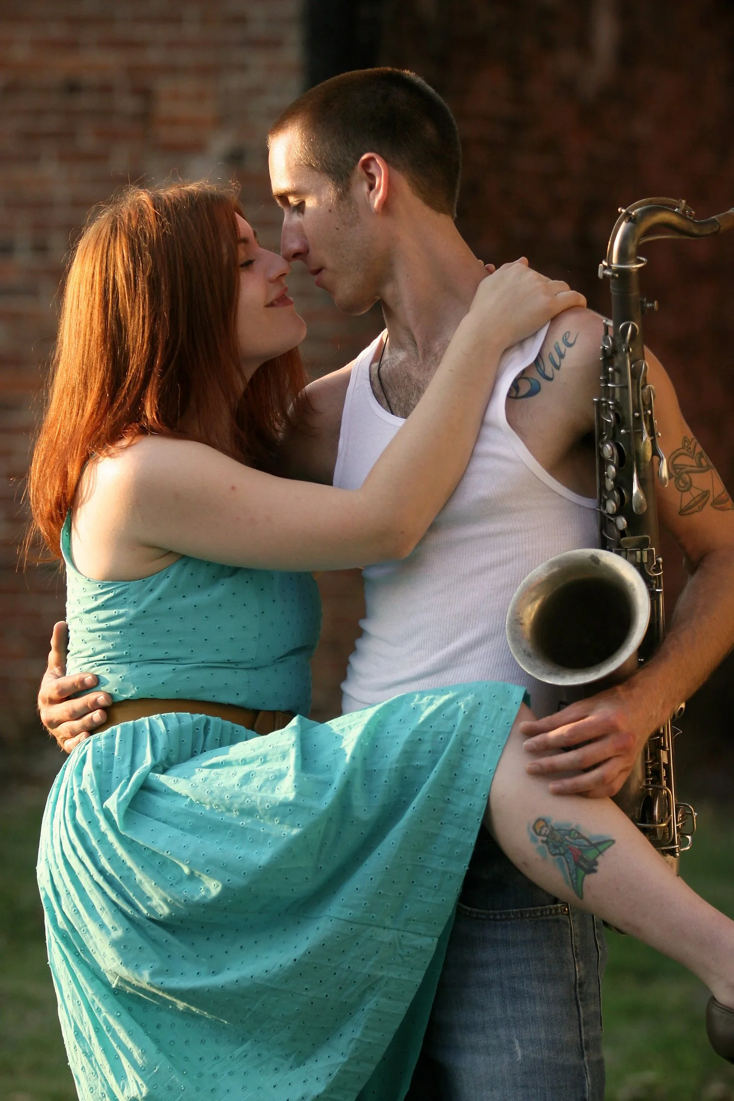 Vertical photograph of a couple taken with late-day golden light. She has red hair and is wearing a turquoise dress. Her right arm is over his shoulder and her right leg is hiked up around his waist. He is wearing a white tank top and jeans and holdi