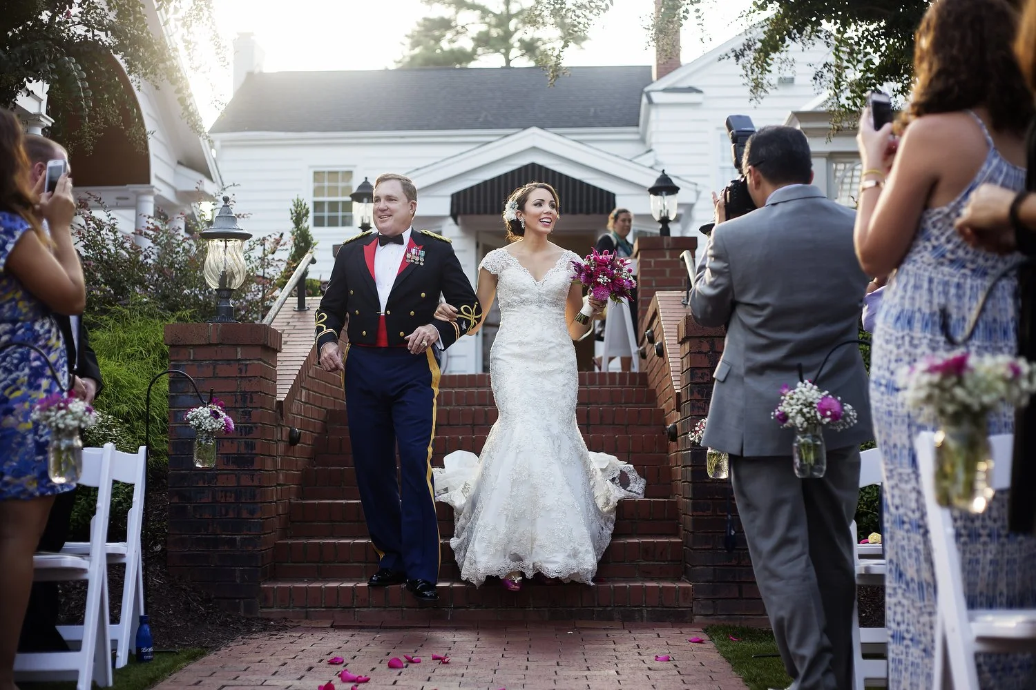 Bride in a white wedding gown and her father in a military uniform are walking down outdoor brick steps, surrounded by guests taking photos and holding floral arrangements, during a wedding ceremony in the late afternoon.