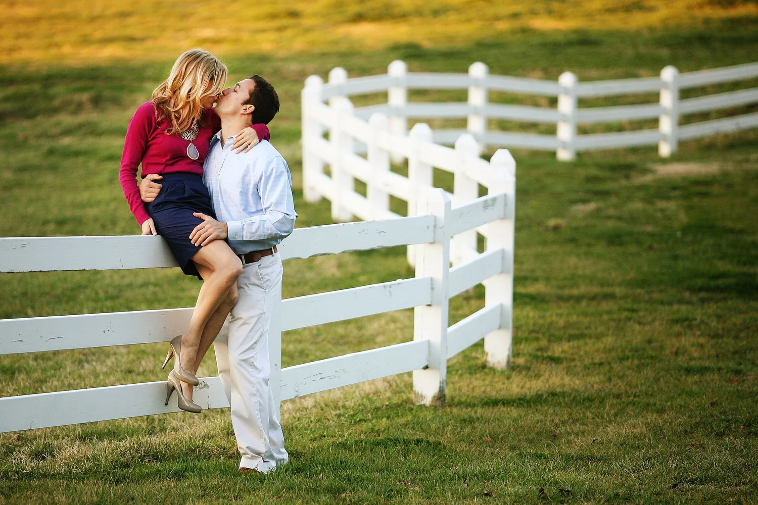 A woman in a red long-sleeve shirt and short skirt and high heels is sitting up on a white wooden fence. Her shapely legs are being held at the thigh by a man kissing her. He stands next to her in khaki pants and a pale blue dress shirt. The wooden f
