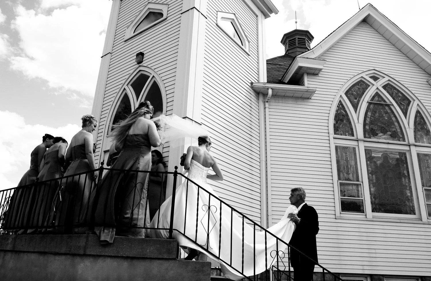 A bride in a wedding gown with her back to the camera waits on an outdoor staircase with a wrought iron railing leading up to an old white wooden church with arch-shaped stained glass windows. Her veil is blowing in the wind and she has all of her br