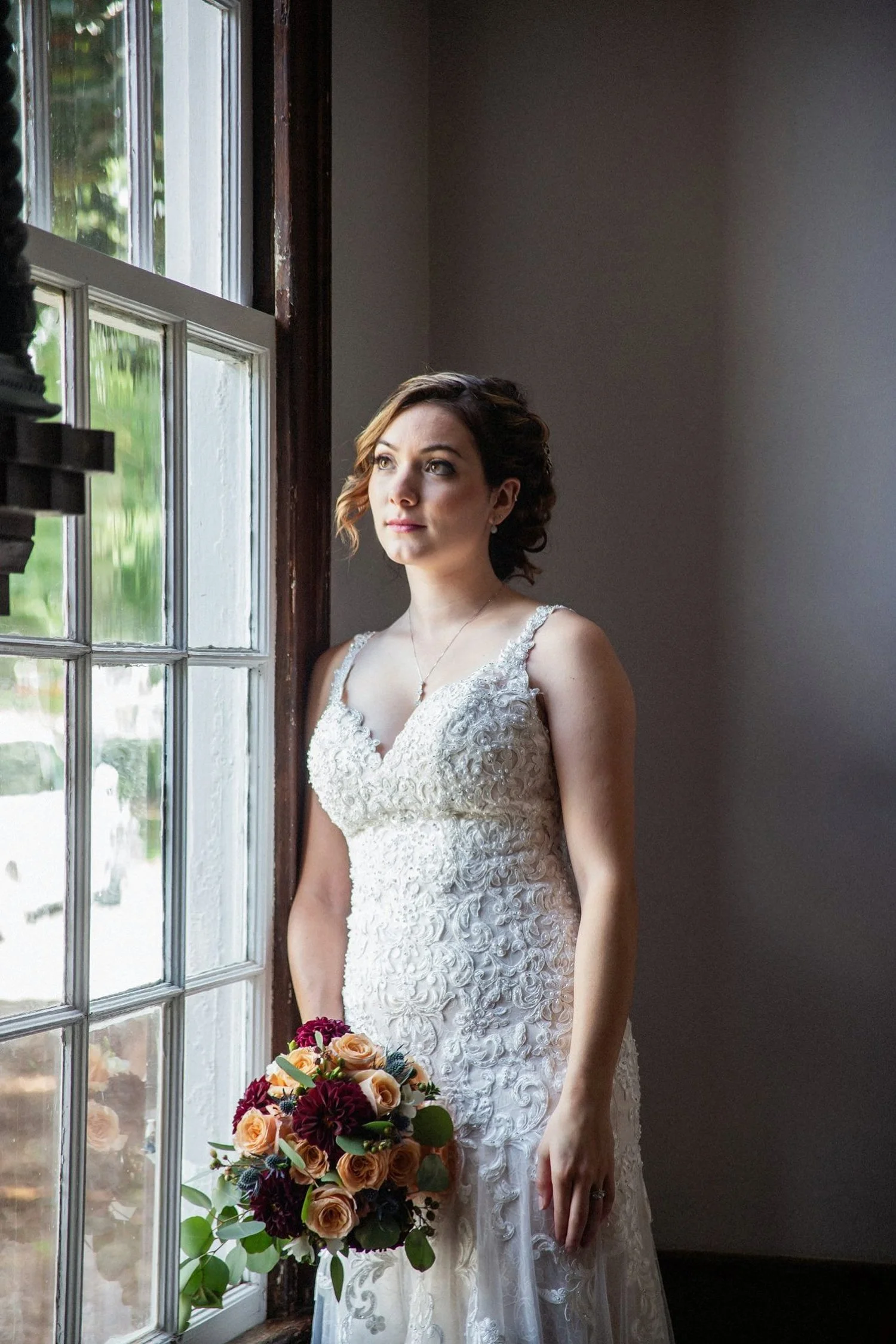 A bride in a lace wedding dress holding a bouquet of flowers stands by a window, looking outside contemplatively.