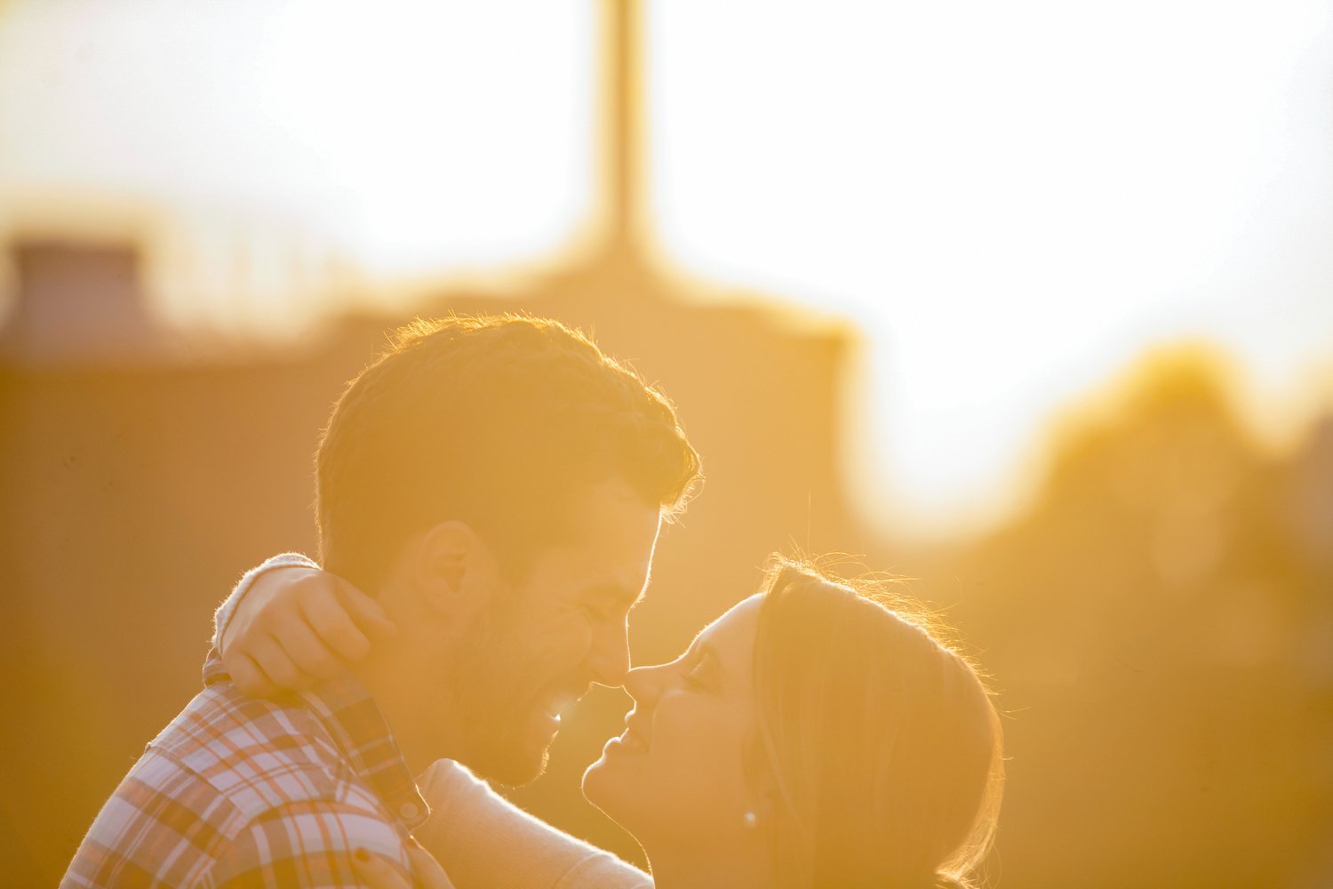Late day warm sunlight glow frames a couple as they face each other and touch noses while smiling. Her hands are wrapped around his neck, and he is wearing a checkered print shirt. It is a close up image with just their heads and a small part of thei