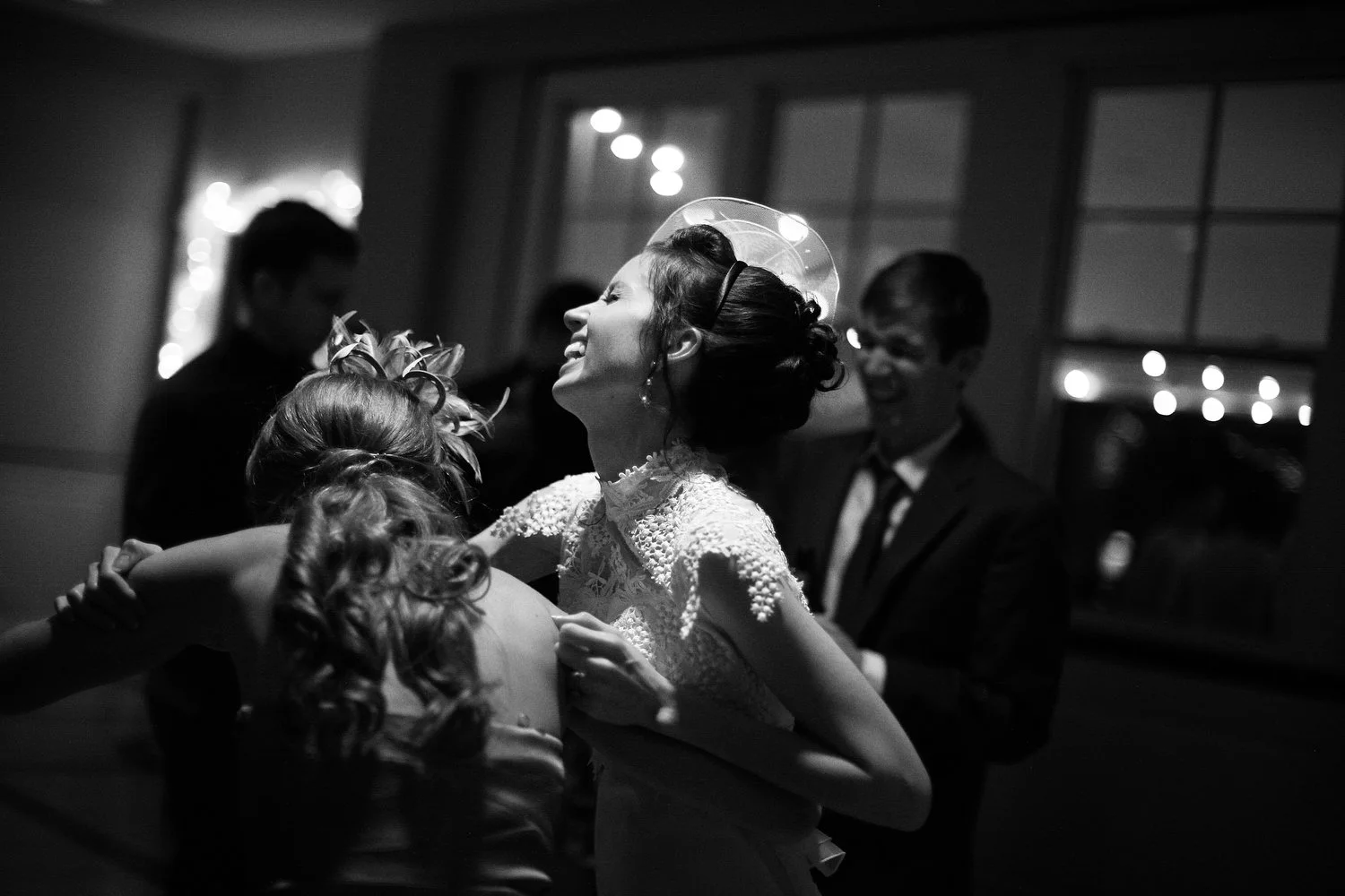 Tight black and white photo of a bride in a vintage white dress looking up and grinning. Her dark hair is adorned with a fascinator. The doors with windows behind her show lights out in the distance, perhaps a city behind the reception hall. A few gu
