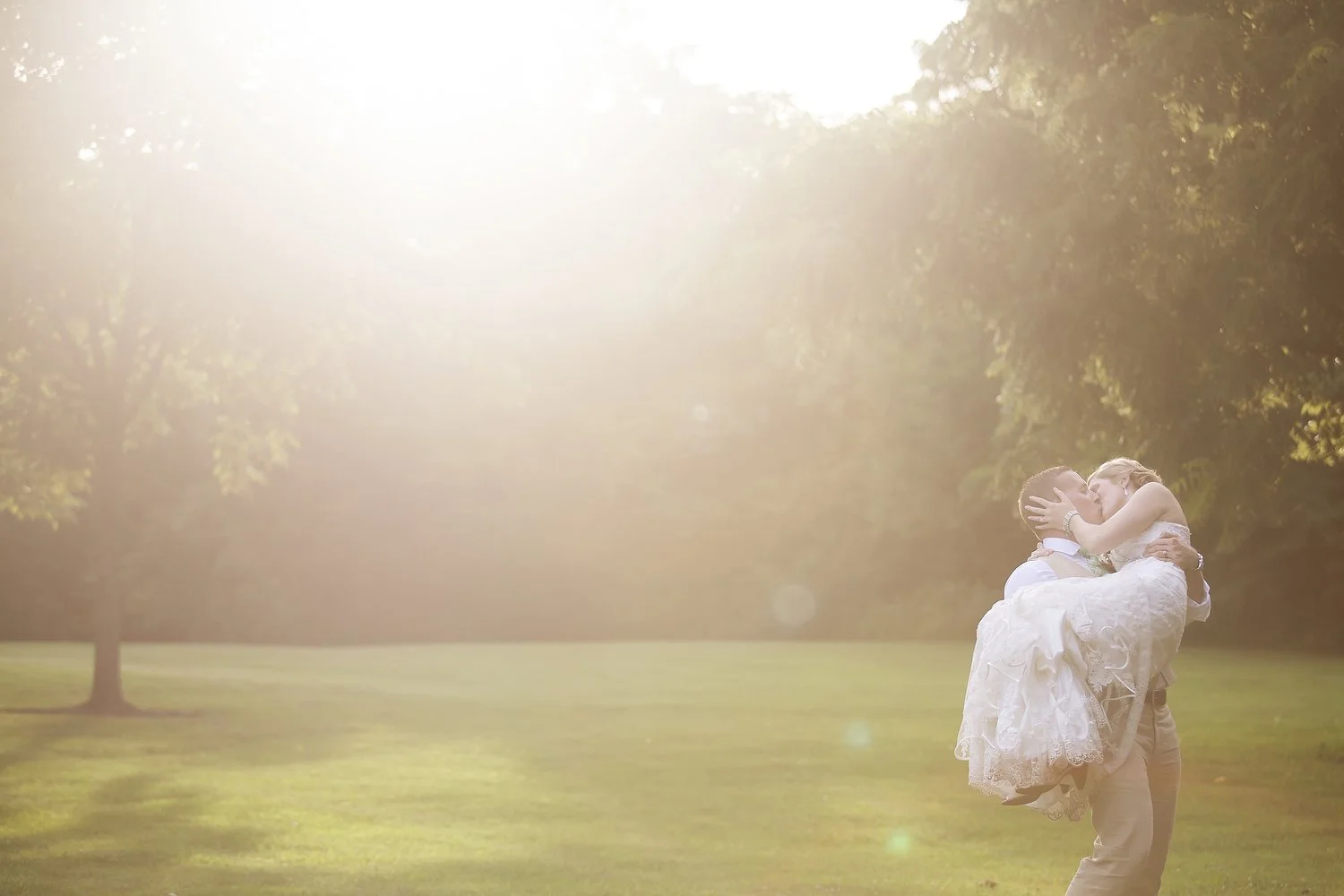 Golden sunlight at sunset swirls around a bride and groom in a green grassy field with trees all around. He is in a tan suit and holding her up in his arm cradling and kissing her. She has on a. vintage white dress and clutches his face in her hands.