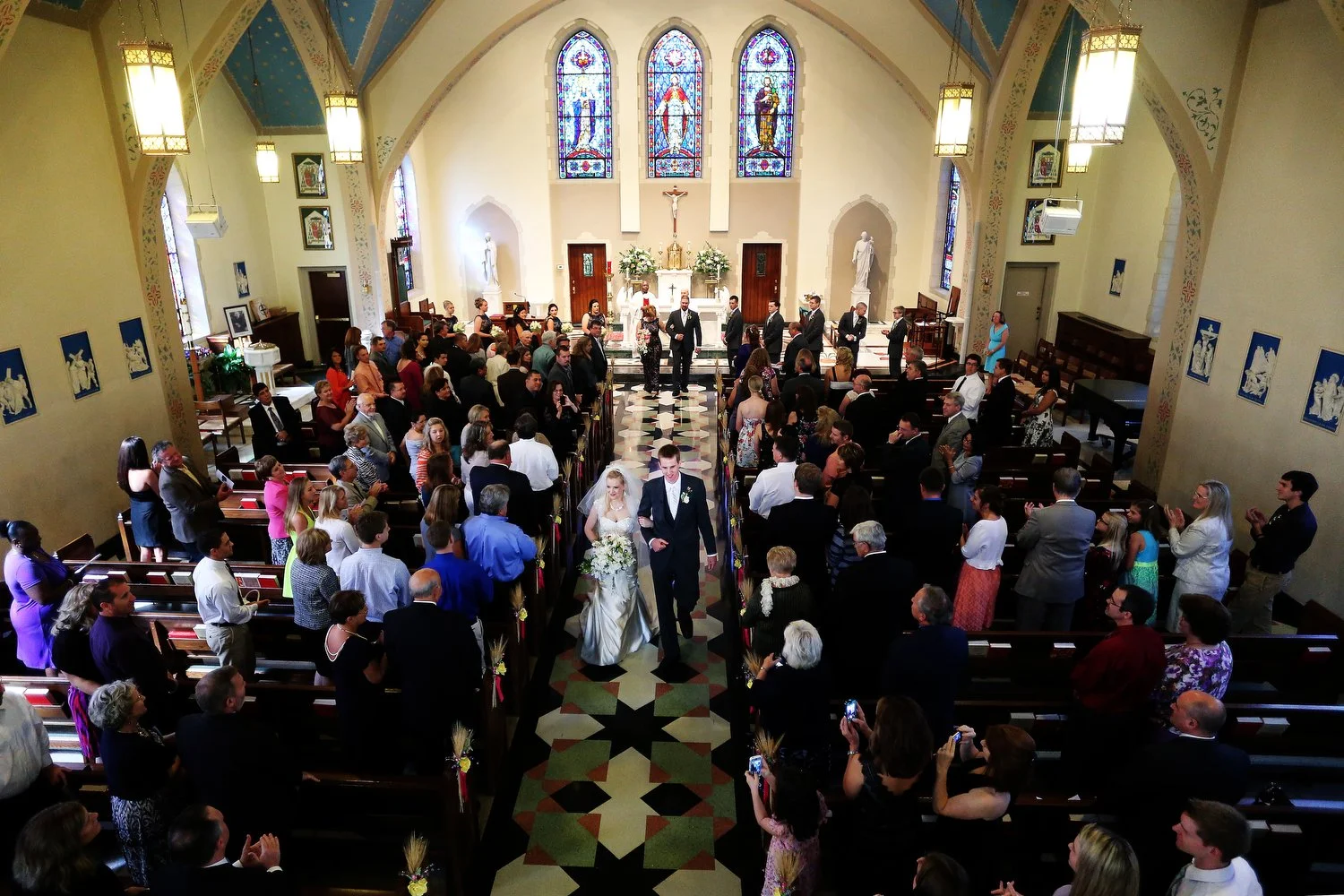 Wedding ceremony in a church, bride and groom walking down the aisle, surrounded by guests and the wedding party, with stained glass windows and religious statues in the background. The checkerboard tile floor is seen from an above angle.