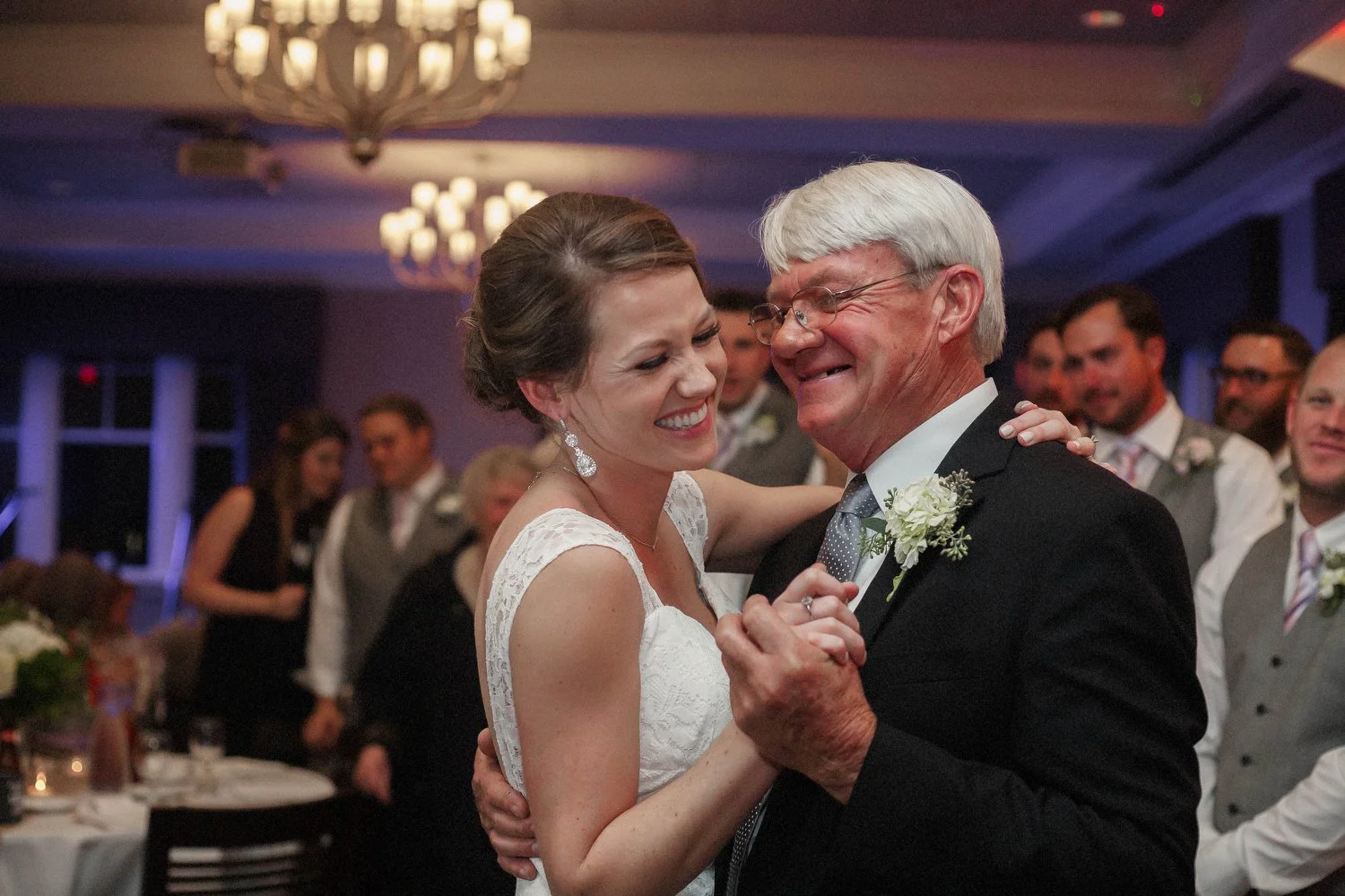 A bride and an older man, likely her father, sharing a dance at a wedding reception in a ballroom with chandeliers and guests in the background.