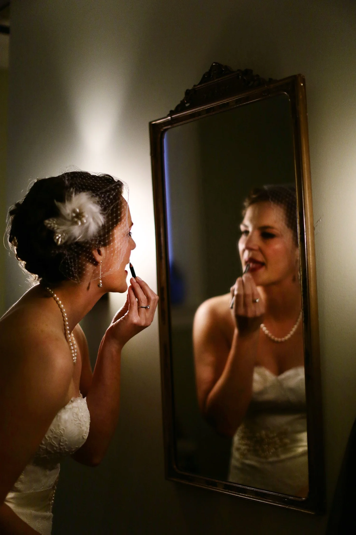 A woman in a strapless wedding dress wearing a white birdcage veil and pearl necklace applies lipstick while looking in a mirror in a dimly-lit room with wall sconces.