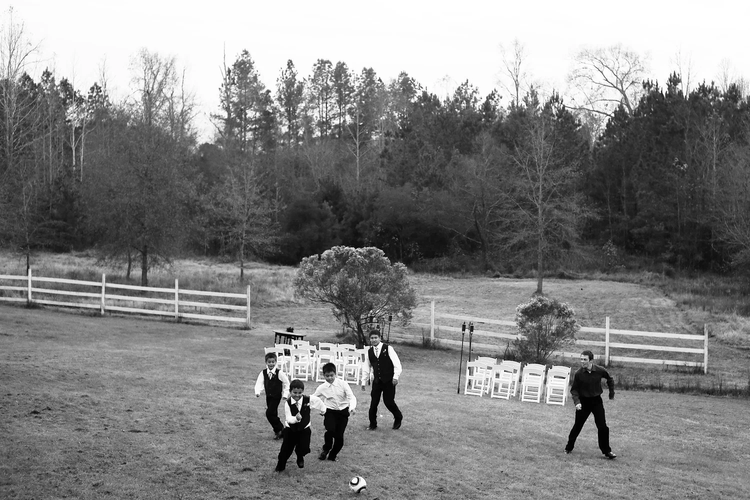 A group of boys playing soccer on a grassy field outdoors with a backdrop of trees, chairs, and a white fence.