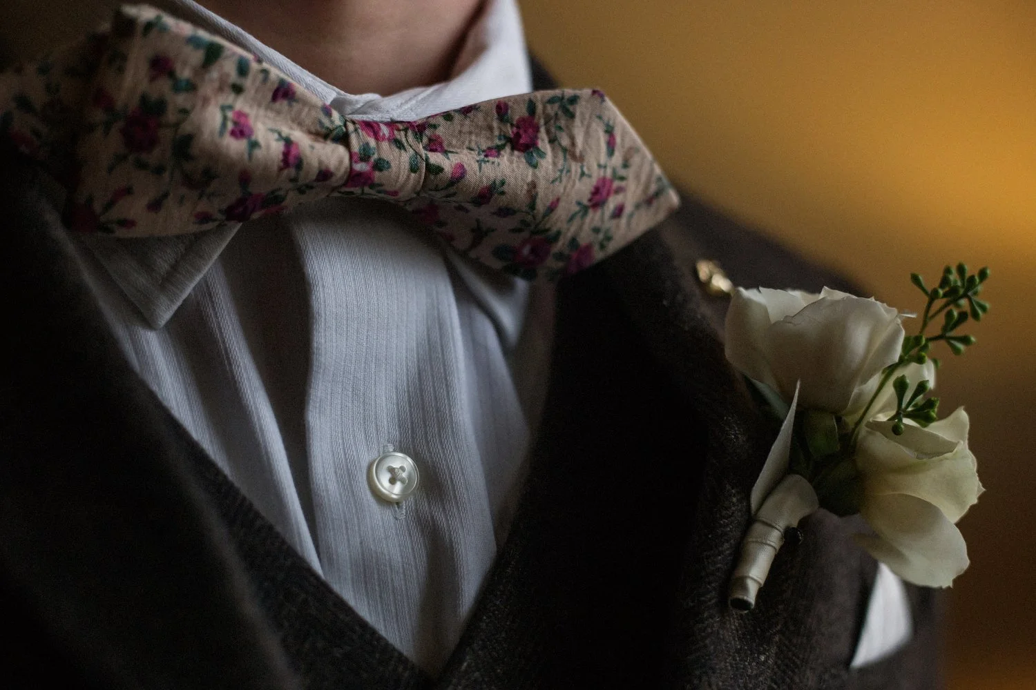 Close-up of a man in a dark suit jacket wearing a floral-patterned bow tie, a white dress shirt, and a boutonniere with white flowers and greenery on his lapel.