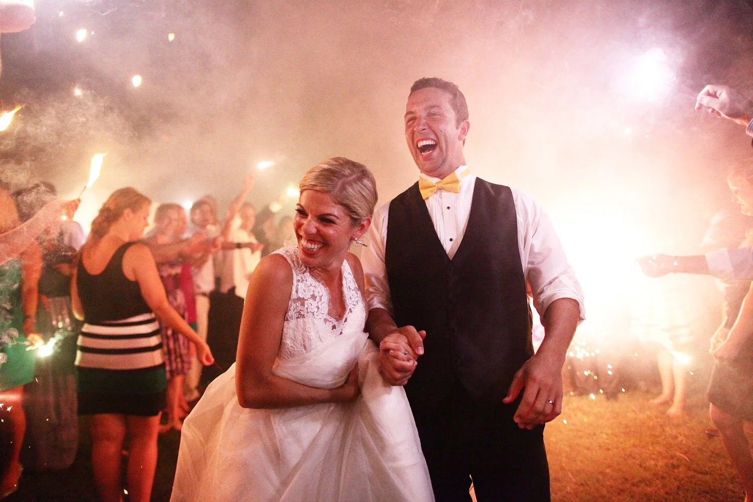 A bride and groom laughing and holding hands at their wedding reception with guests in the background holding sparklers.