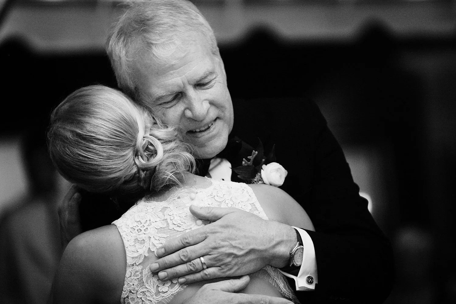 Close black and white view of an older man smiling and leaning in to touch his head against his daughter's head as they embrace at the wedding reception. His hands are around her on her back and only his face is shown. He is wearing a black tuxedo an