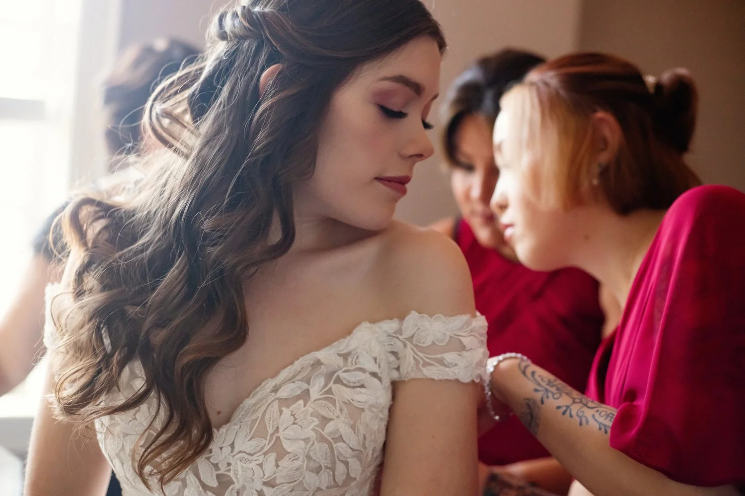 A woman looks over her shoulder as women behind her help fasten her white lacy wedding dress.