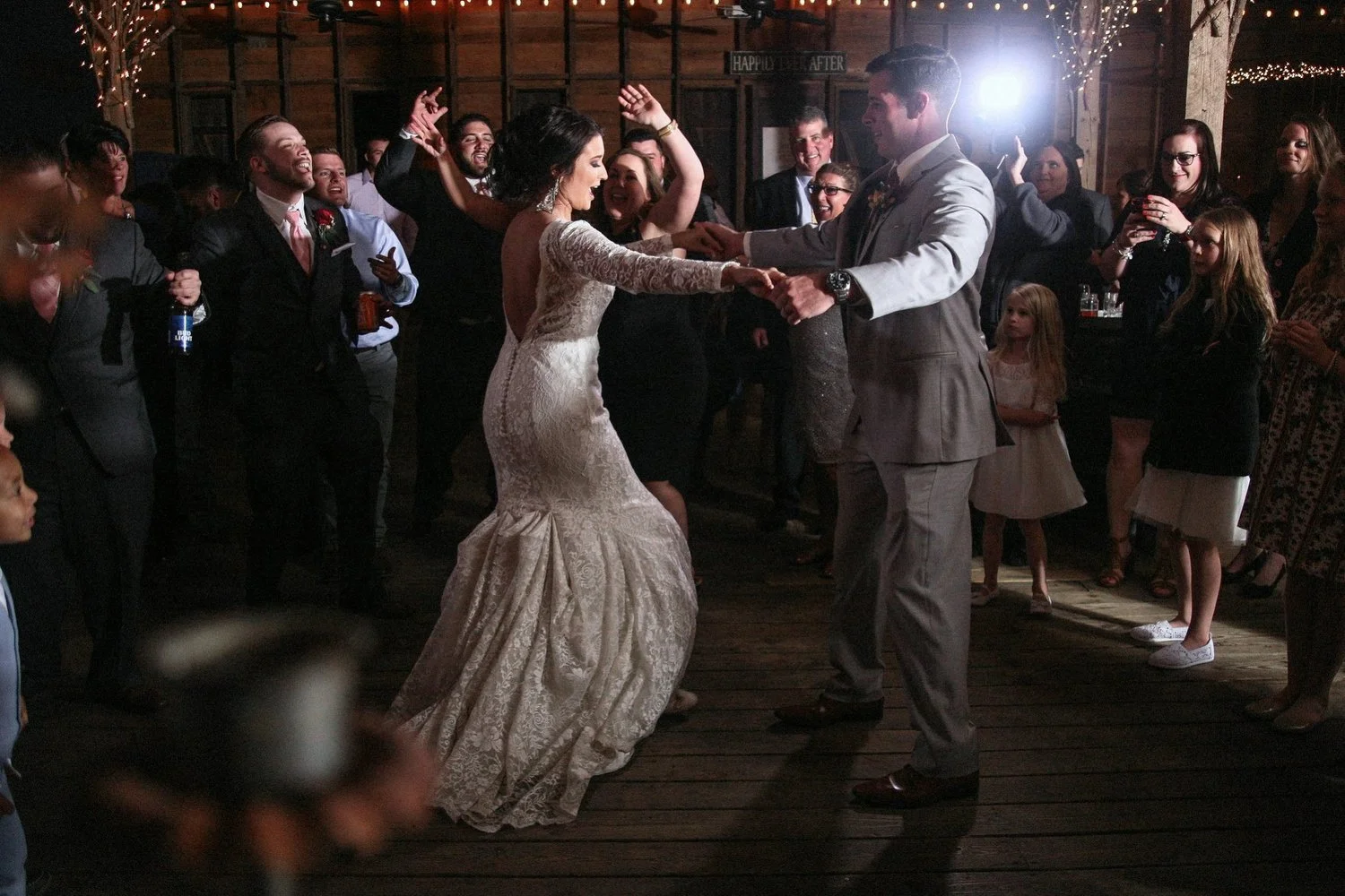 A bride and groom dance at their wedding reception surrounded by friends and family. She is wearing a vintage dress with long sleeves, and an open back that is tight-fitting until it reaches just above her knee and then flows outward at the bottom. H