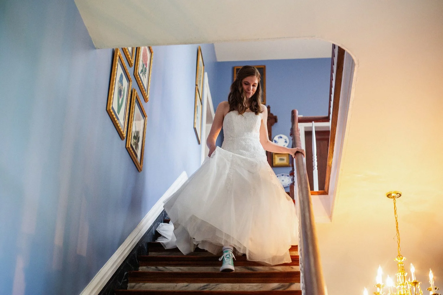 A young woman holds her white wedding dress up as she walks past blue walls down a staircase. Her converse shoes are visible.
