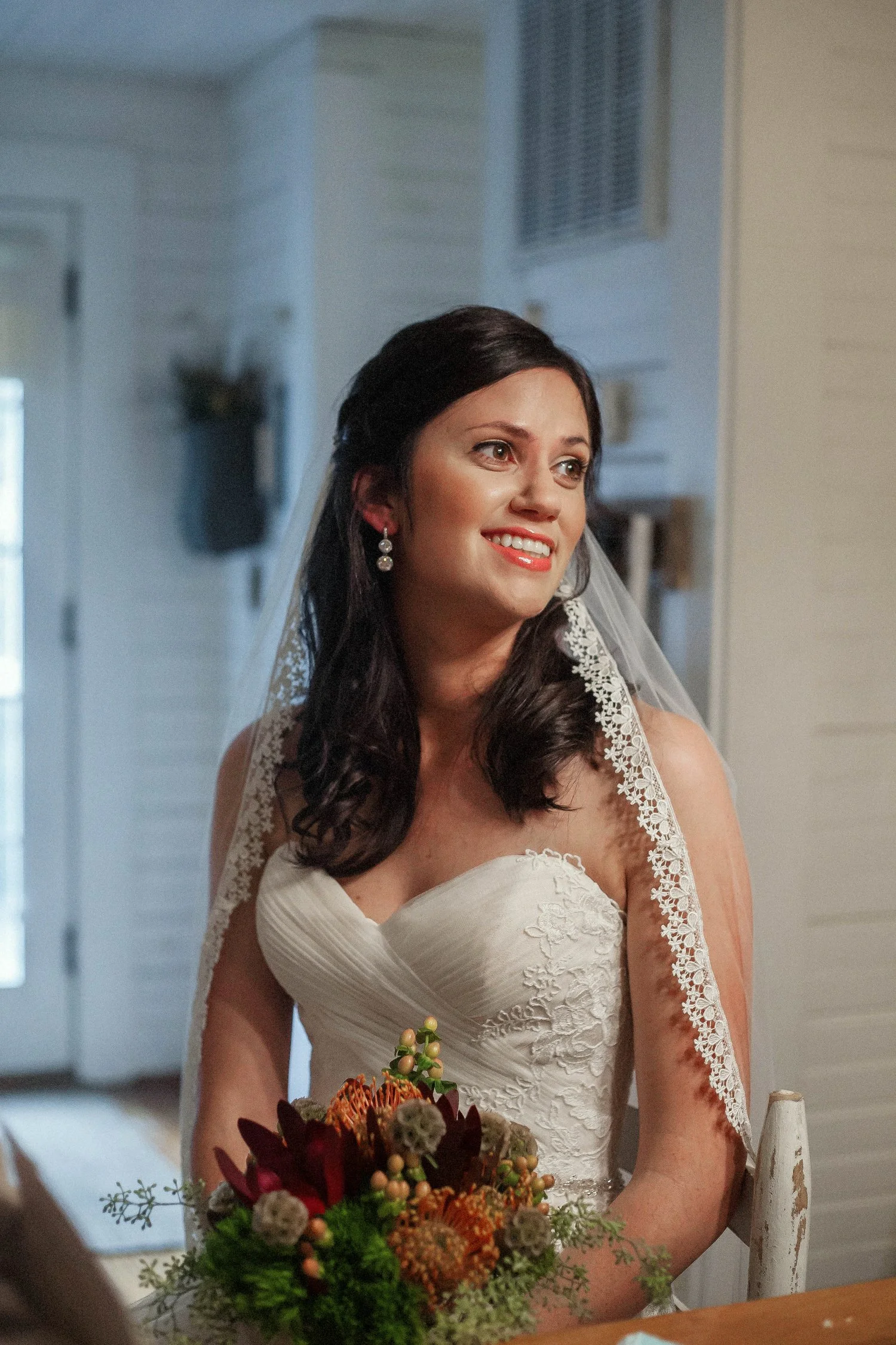 A bride in a strapless white lace wedding dress, with dark hair and earrings and a veil, smiling while holding a bouquet of flowers in an indoor setting.