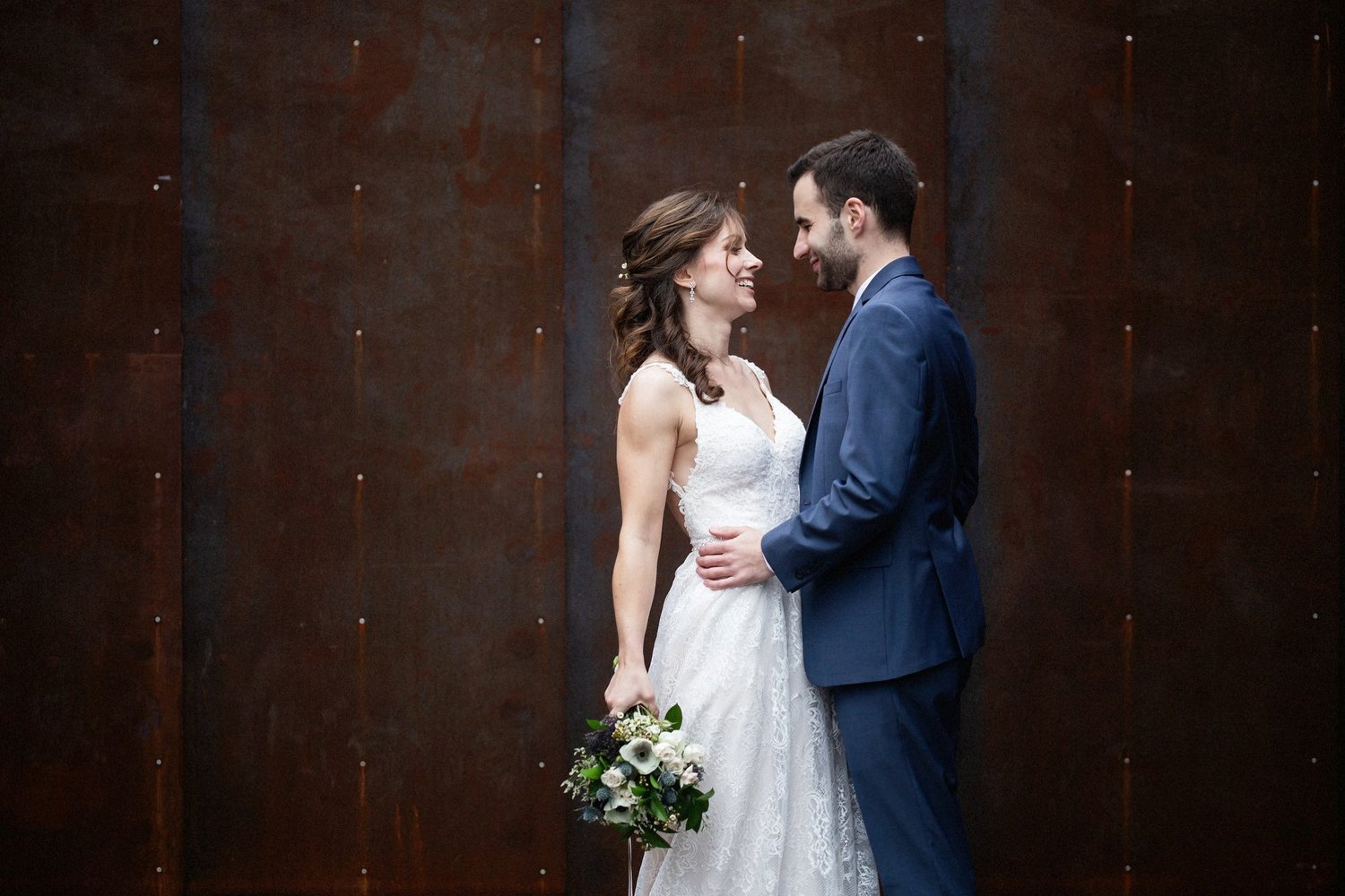A bride and groom standing close together, smiling at each other, on their wedding day. The bride is holding a bouquet of flowers, wearing a white lace wedding dress, and the groom is in a blue suit. They face each other against a dark, metal wall.
