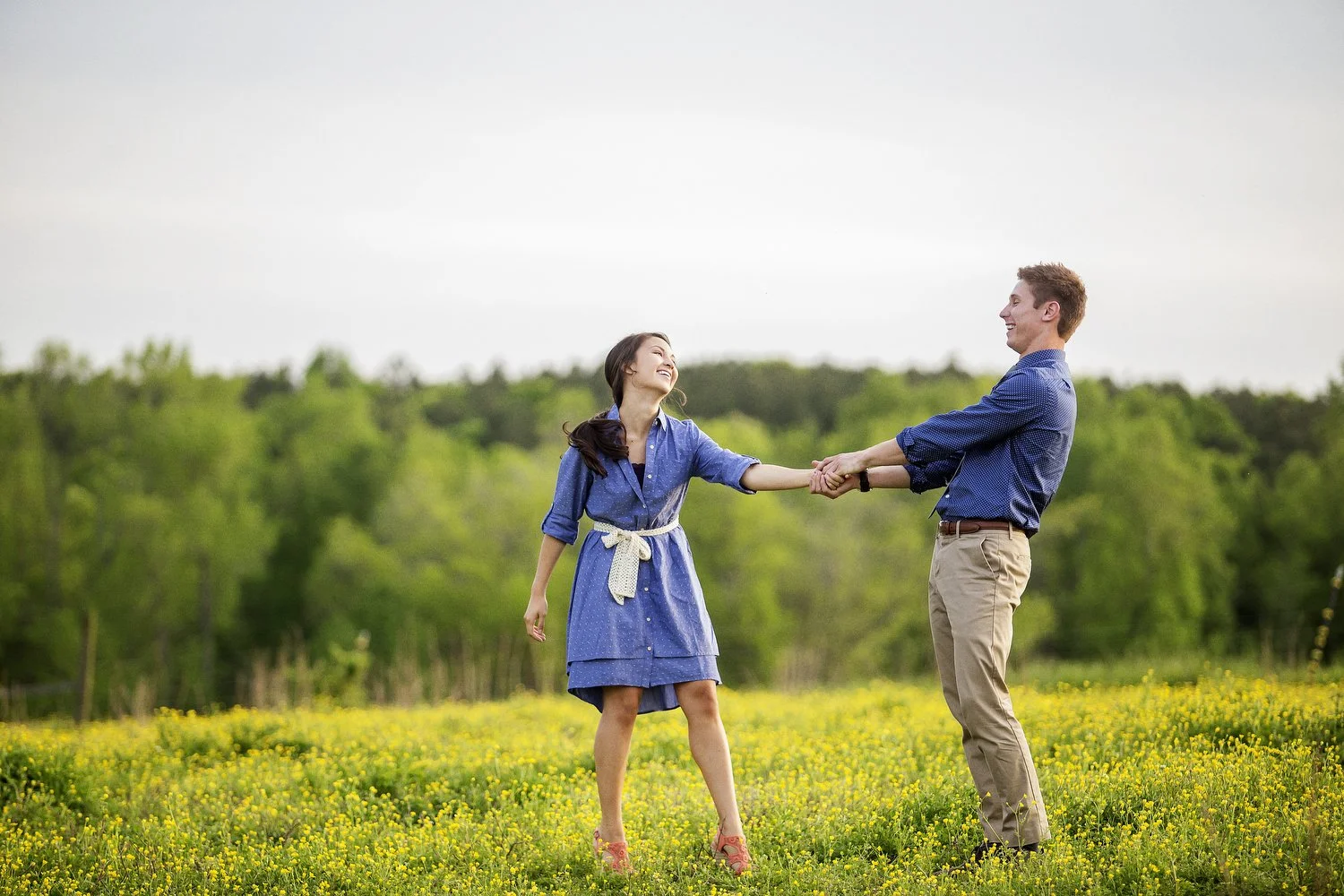 A couple stands in a field of tiny yellow flowers with lush green trees behind them. She is wearing a blue denim dress with a white belt around it. He has on khaki pants and a blue button-up shirt. He is holding his left hand in both of his hands and