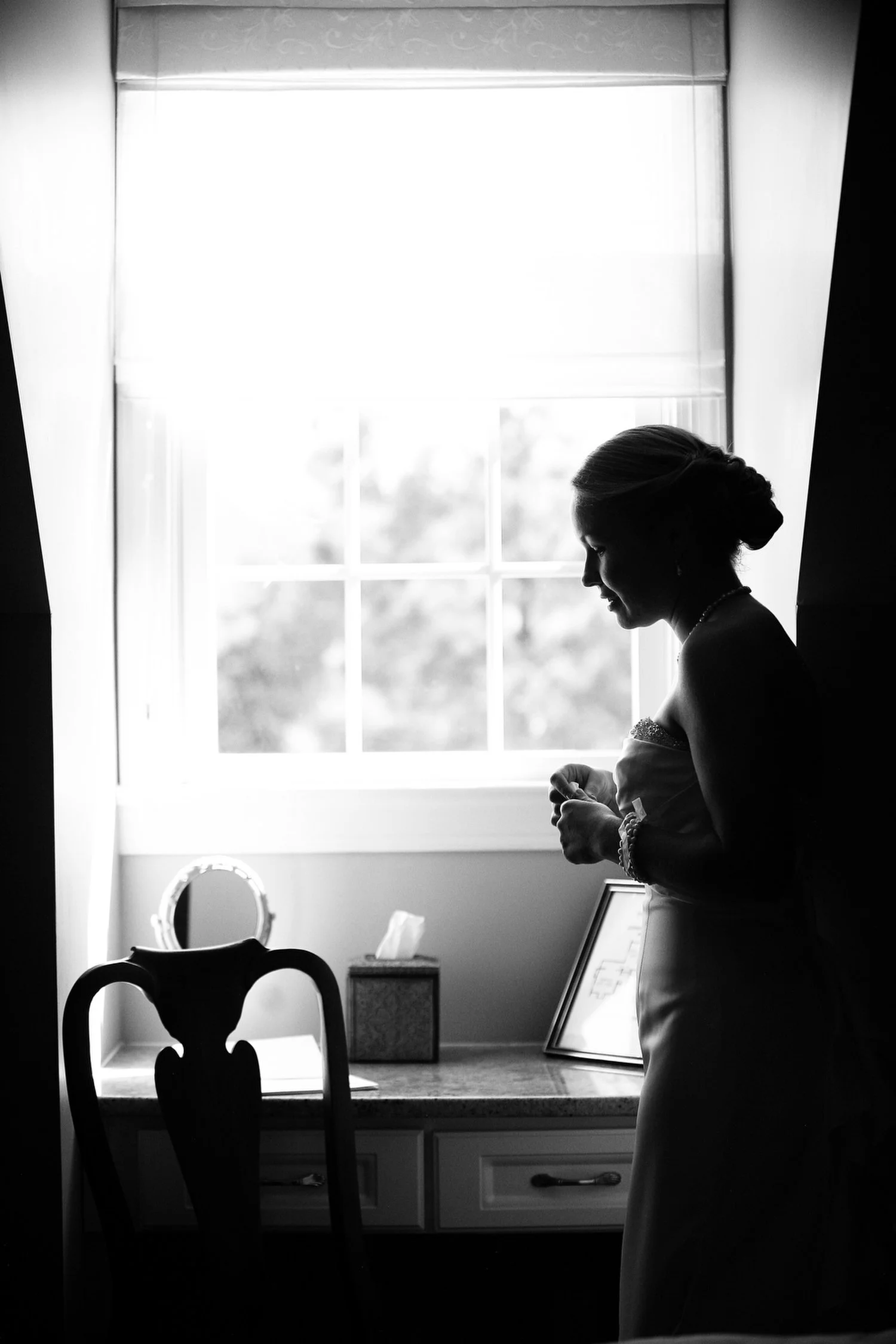 Silhouette of a woman in a wedding dress standing by a window with light behind her, possibly getting ready or reflecting.